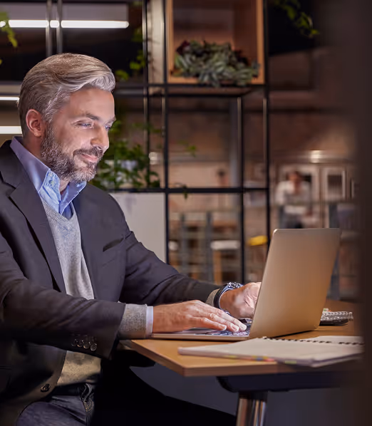 Middle-aged man with gray hair and beard working on a laptop at a wooden desk in a modern office setting.