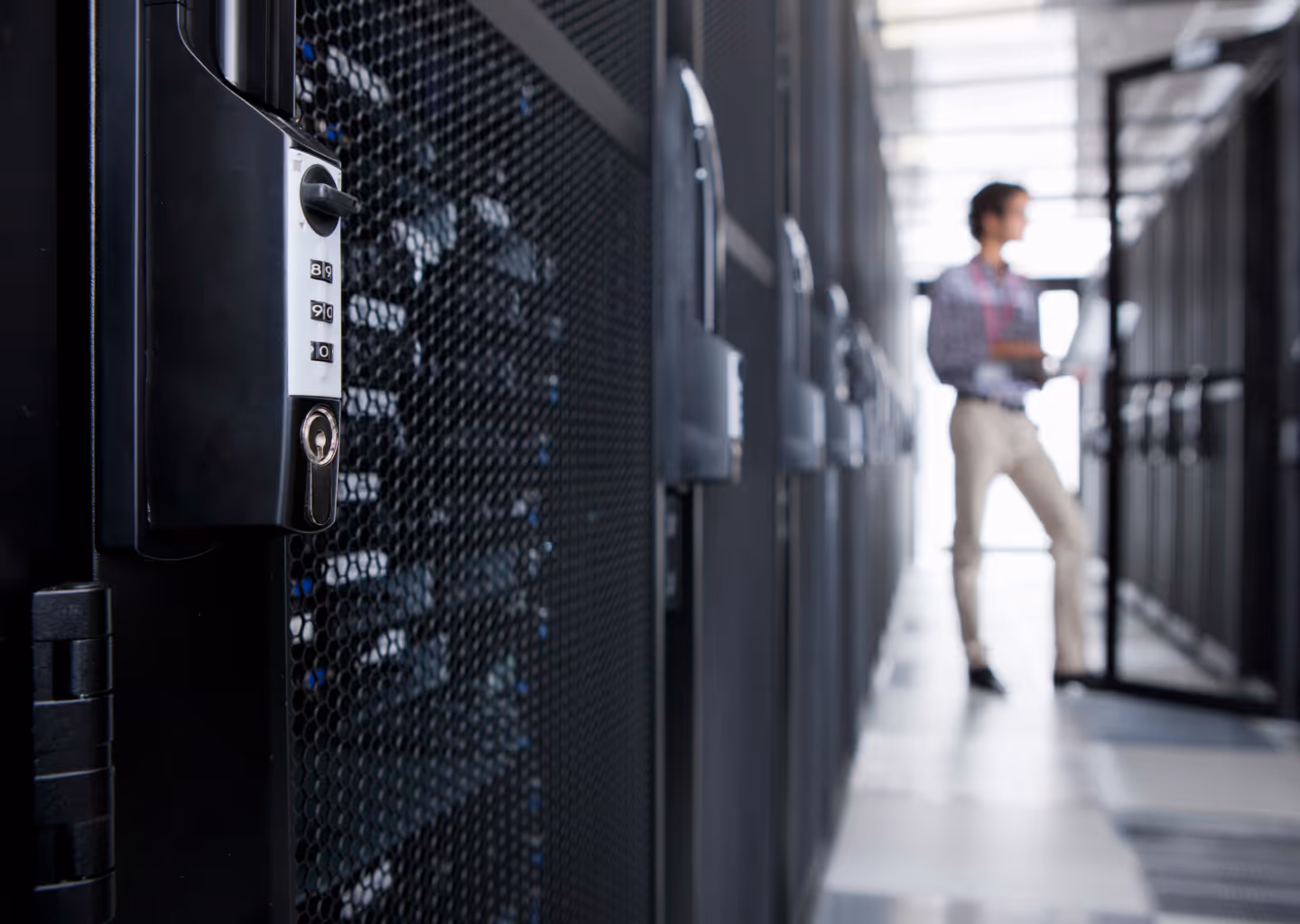 Close-up of a combination lock on a server rack in a data center corridor with a person holding a laptop in the background.