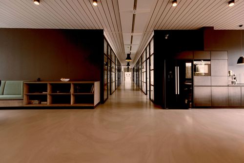 Modern office corridor with glass walls, wood shelving, seating area, and a kitchen with black fridge.