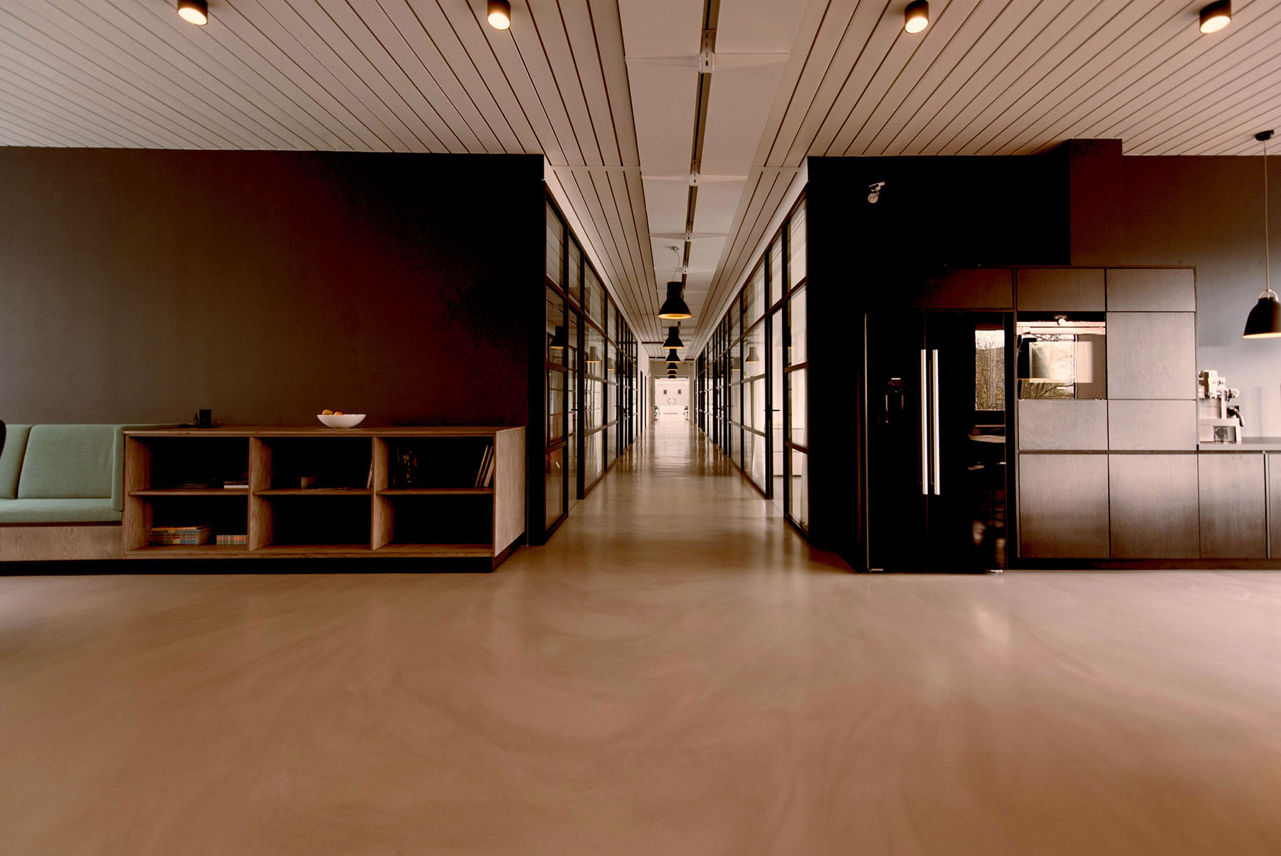 Modern office corridor with glass walls, wood shelving, seating area, and a kitchen with black fridge.