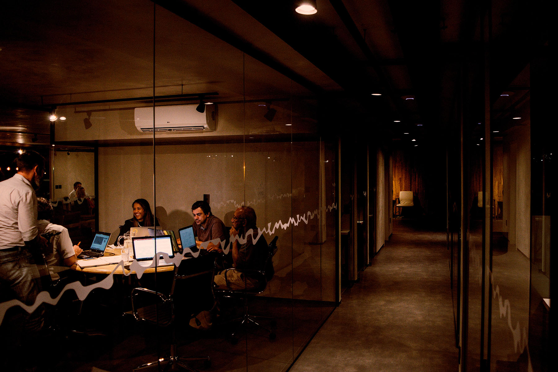Group of people collaborating around a table with laptops in a dimly lit office meeting room.