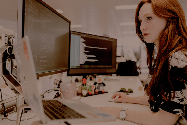 Woman with red hair focused on coding at a desk with dual monitors and a laptop.