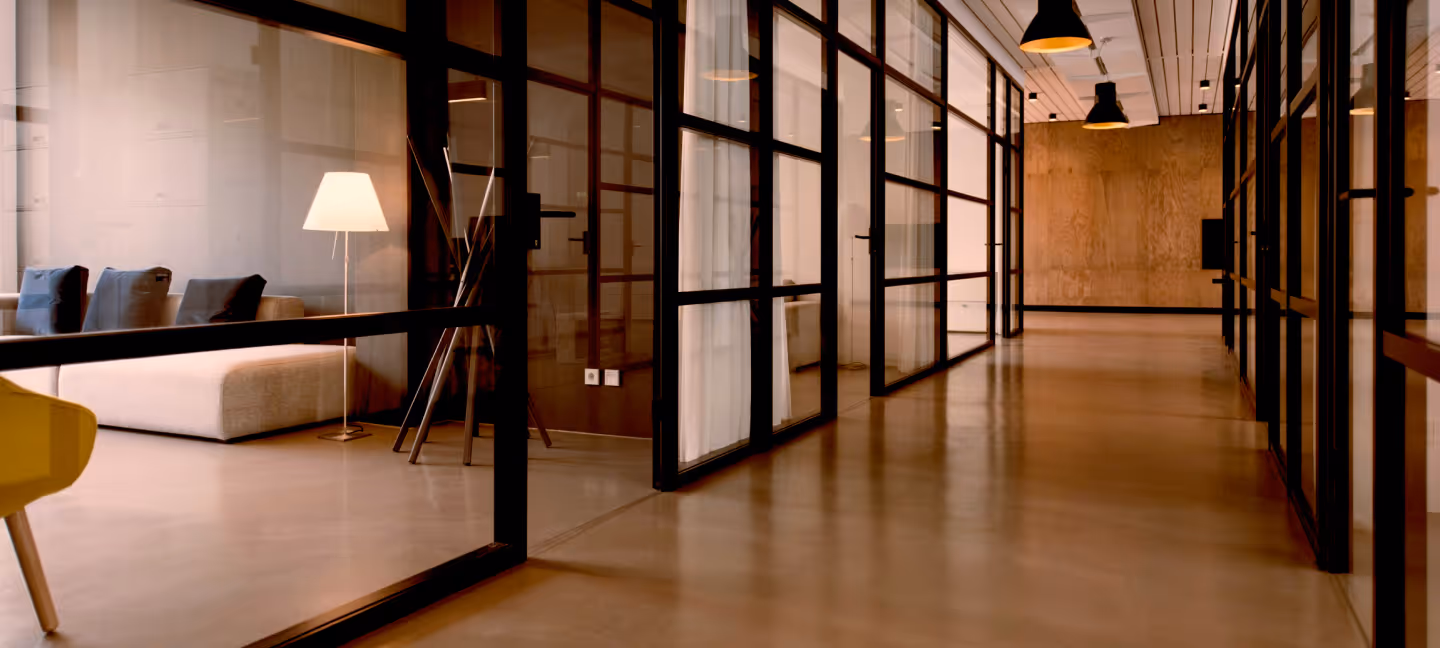 Modern office hallway with glass walls, a beige sofa, floor lamp, and pendant lights.