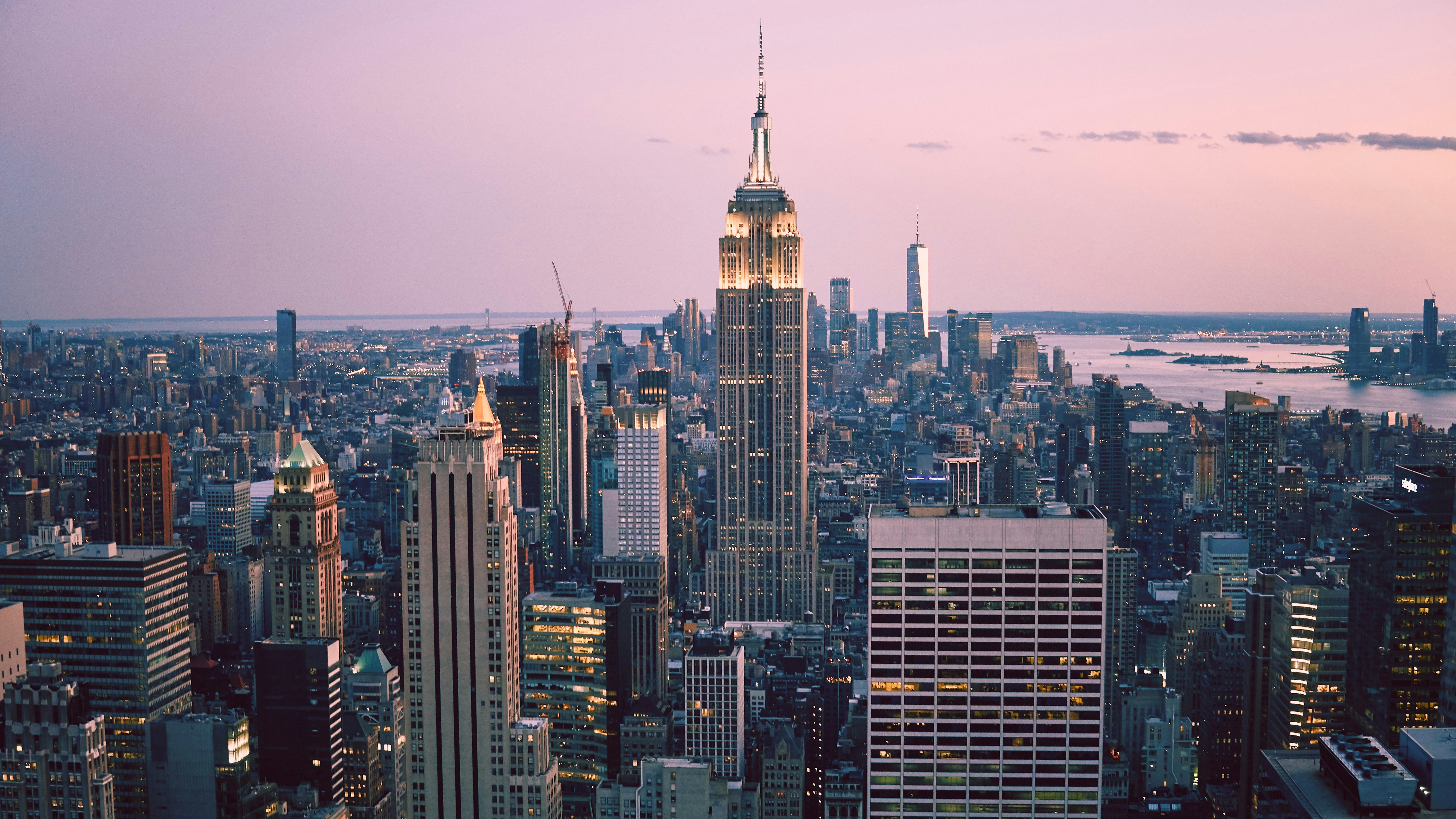 View of New York City skyline at dusk with the Empire State Building prominently lit in the center, surrounded by other skyscrapers and the river in the background under a pinkish sky.