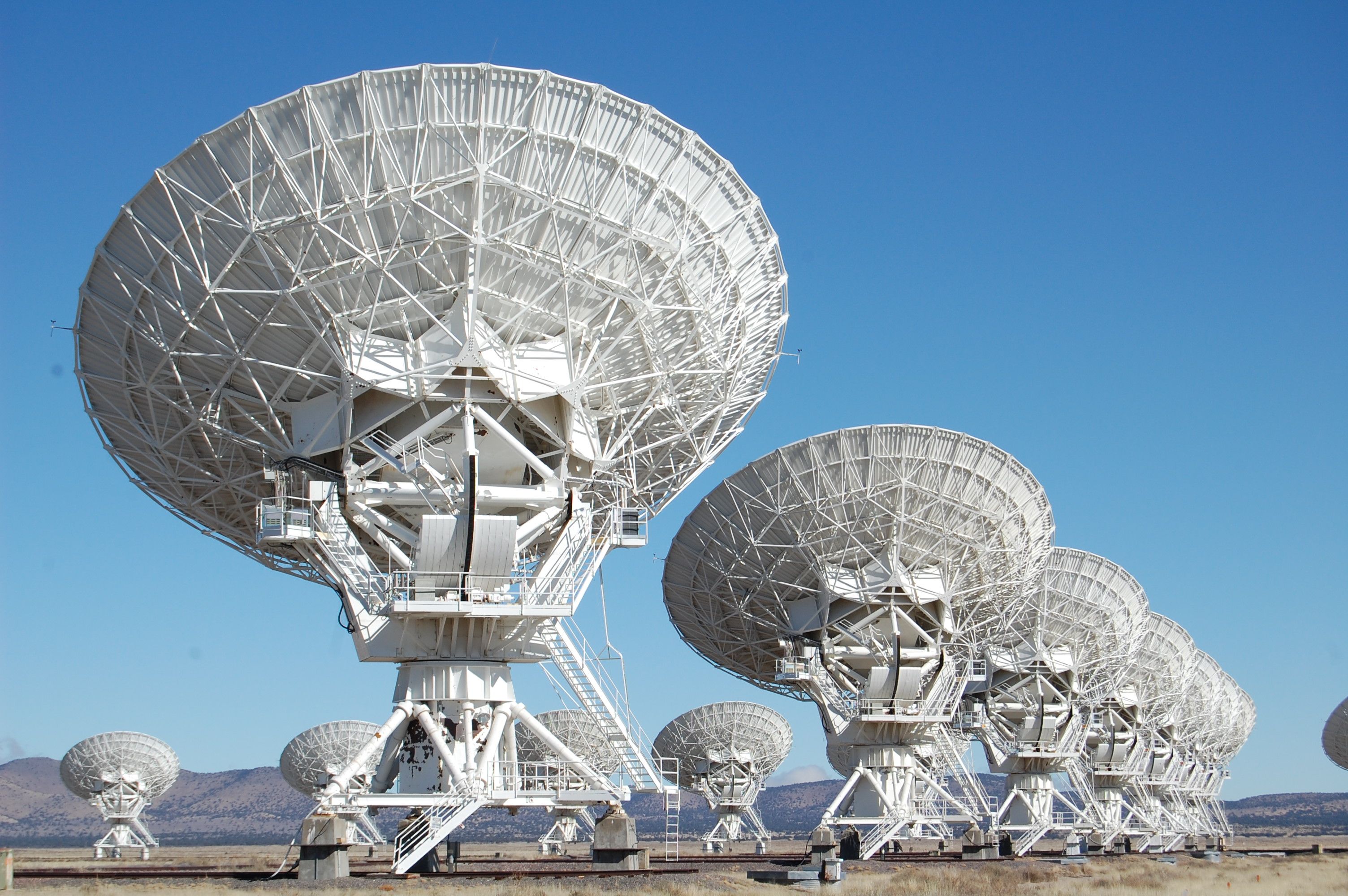 Multiple large white radio telescope dishes arranged in a line on a flat, dry landscape with a clear blue sky and distant mountains.