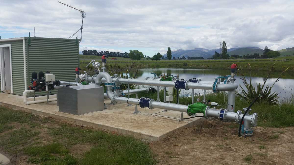 Industrial water pumping station with metal pipes, valves, and a green metal shed near a pond with grassy fields and mountains in the background under a cloudy sky.