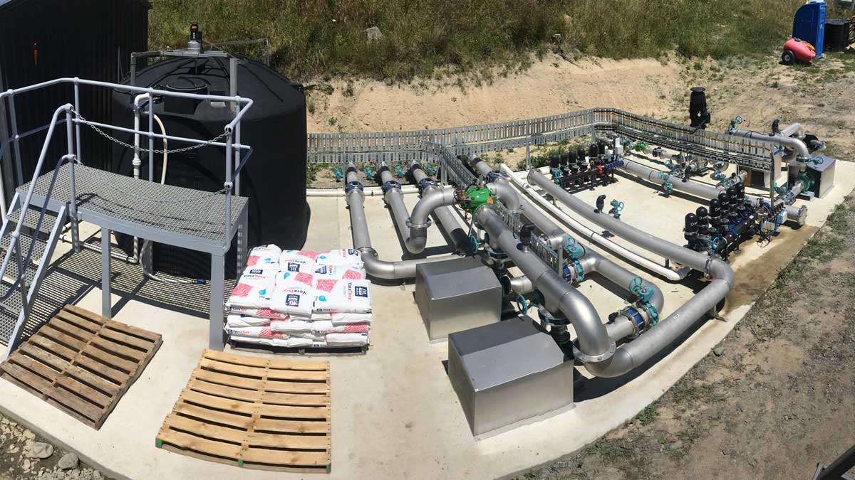 Outdoor industrial water treatment system with large pipes, valves, black tanks, pallets, and bags of chemicals on a concrete pad.