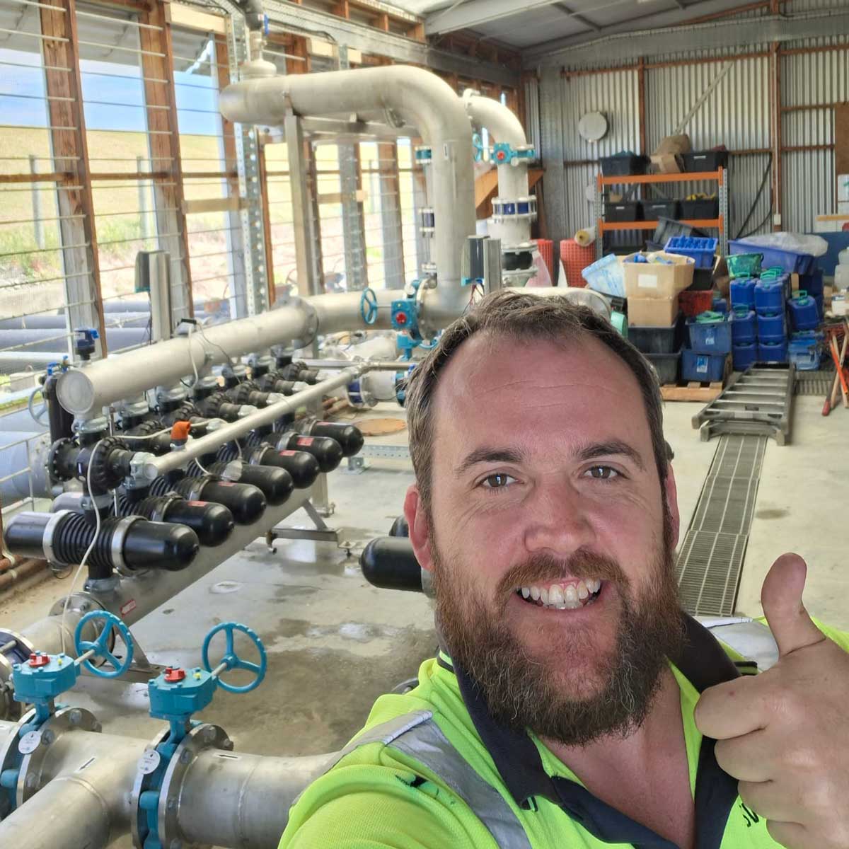 Man in a high-visibility shirt giving a thumbs-up inside an industrial facility with large metal pipes and valves.