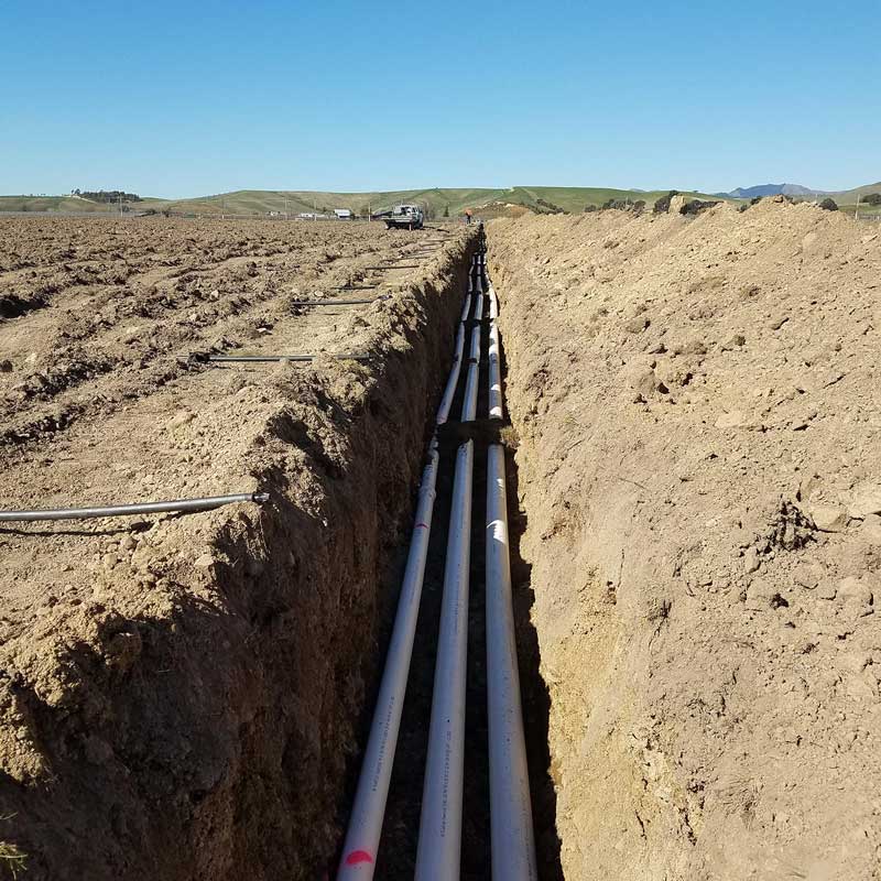 Long trench with multiple white PVC pipes installed at the bottom in an open field under a clear blue sky.
