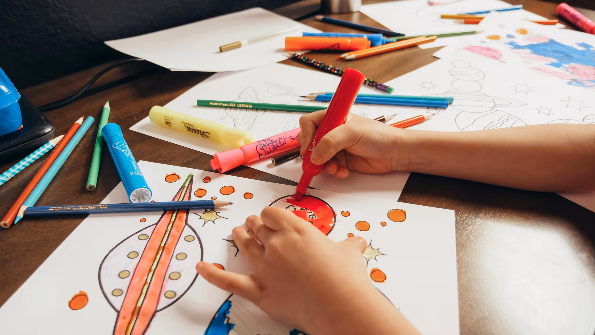 Child coloring a space-themed drawing with markers and colored pencils on a wooden desk.