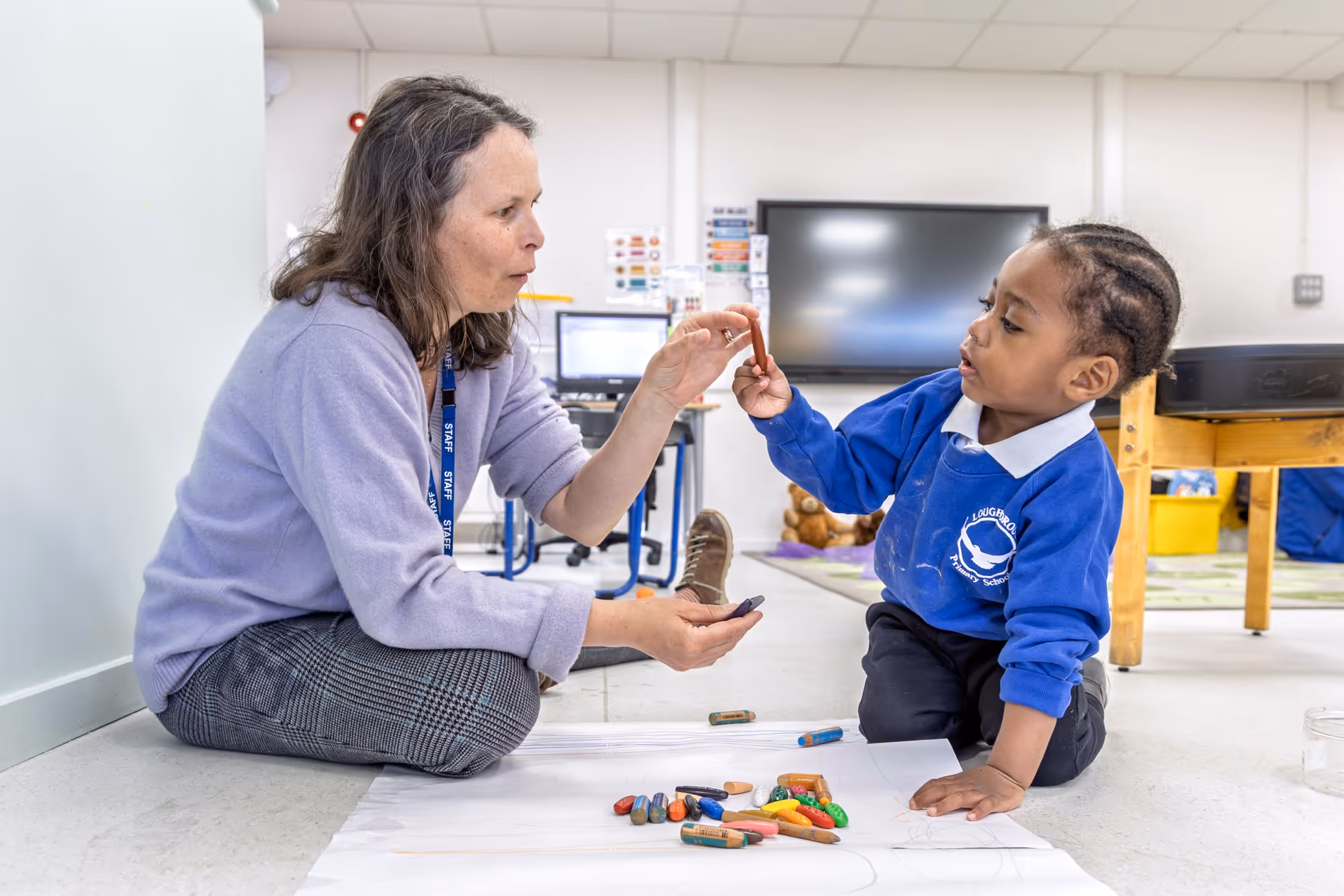 Teacher and child sitting on floor sharing crayons while drawing on large paper in classroom.