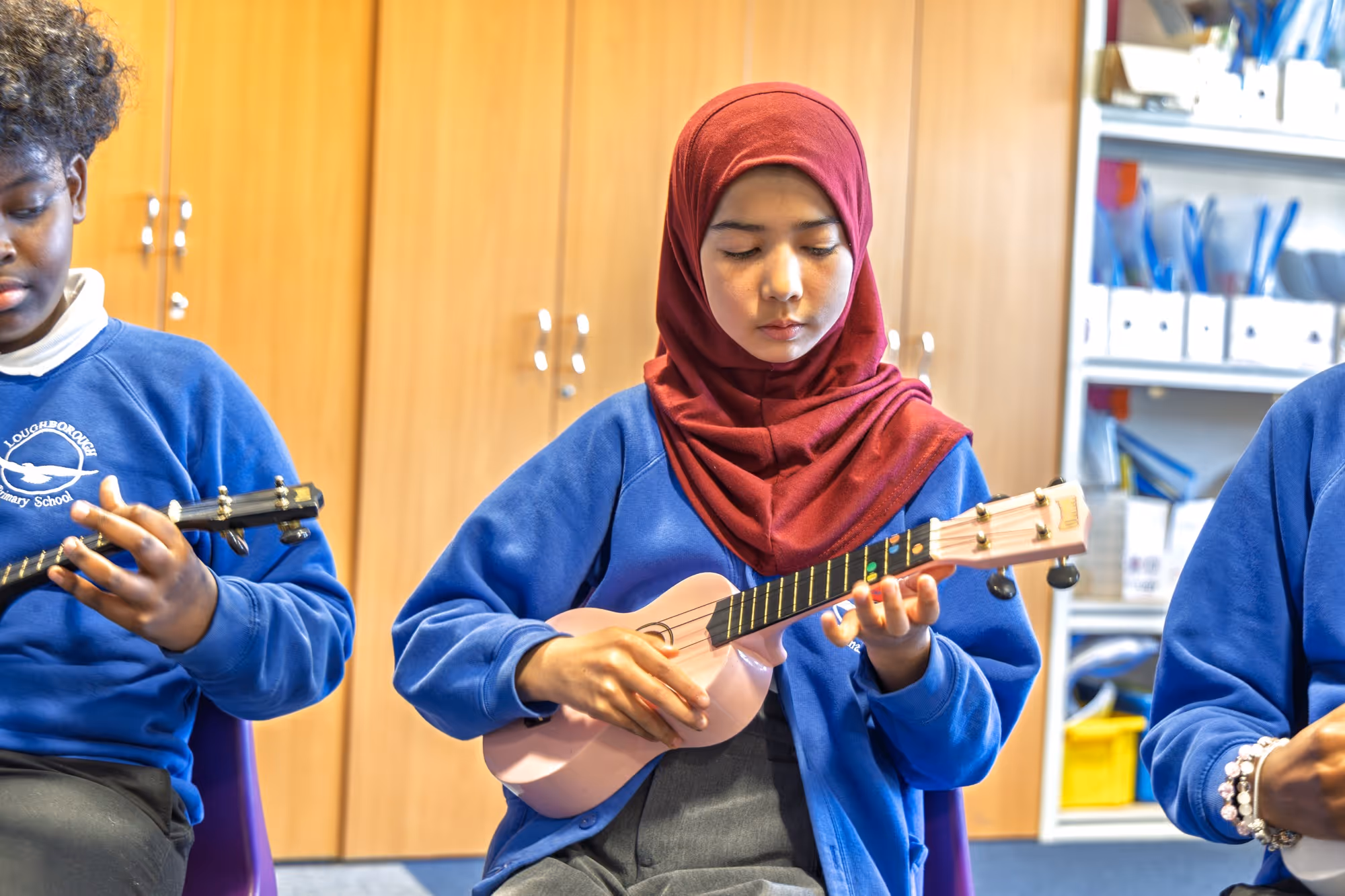 Three students in blue uniforms playing ukuleles in a classroom, one wearing a red hijab.