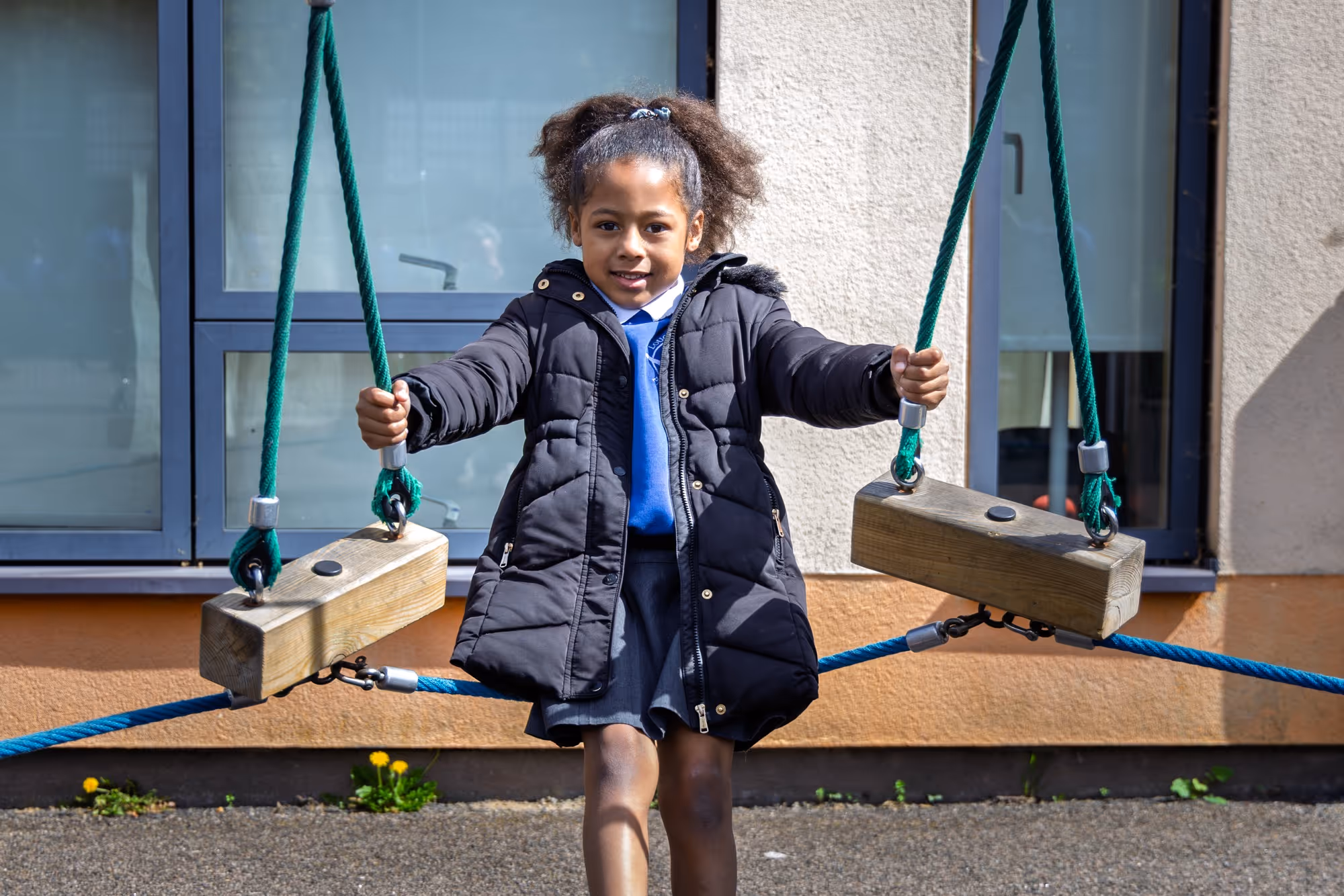 Young girl in a black coat holding ropes while balancing on wooden playground equipment outdoors.