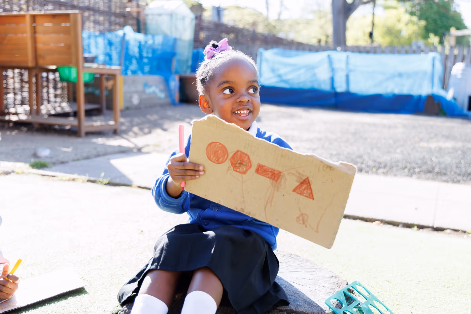 Young girl outdoors holding a cardboard with red shapes drawn on it and a red marker in hand.
