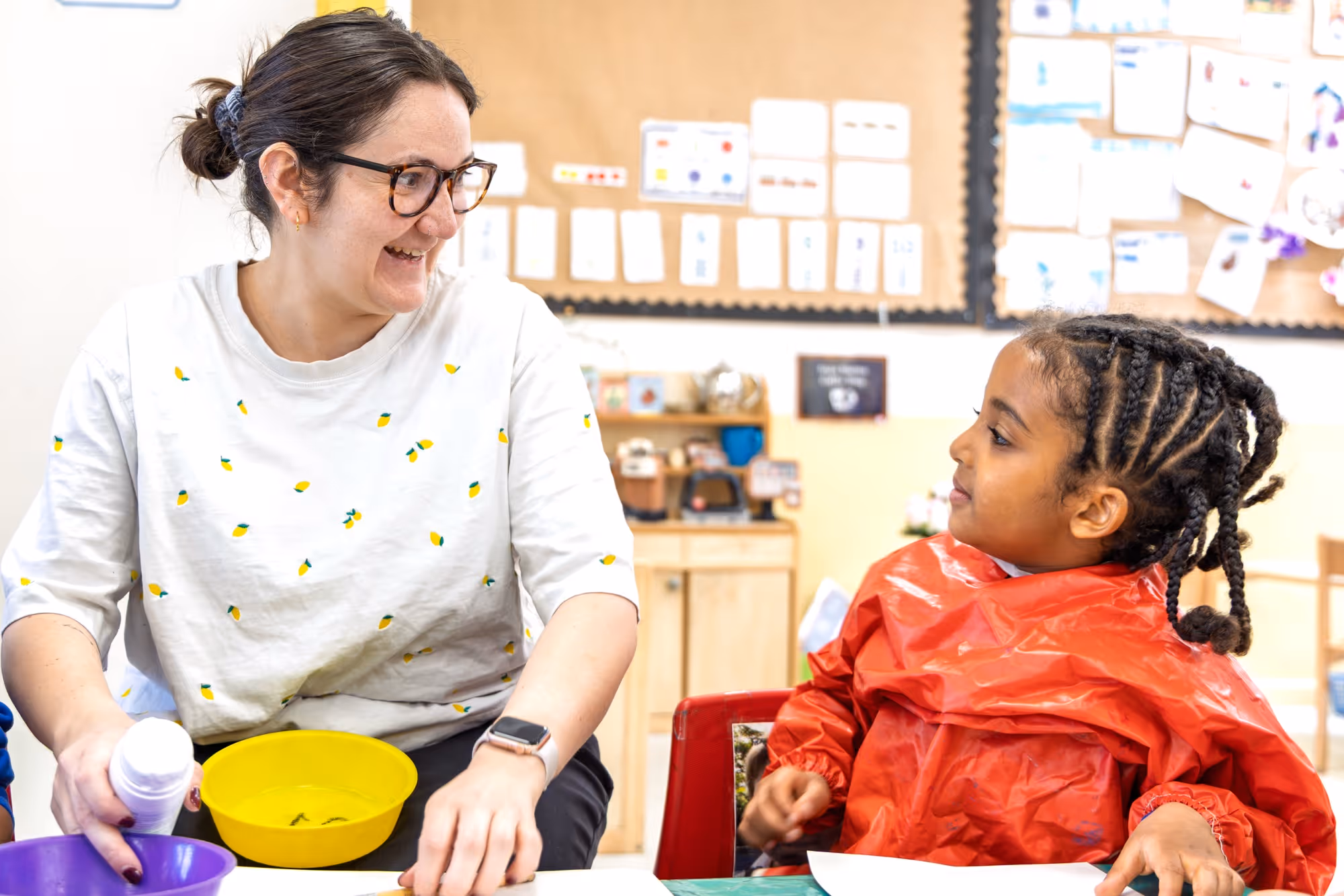 Teacher smiling and engaging with a young girl wearing a red smock in a classroom setting.