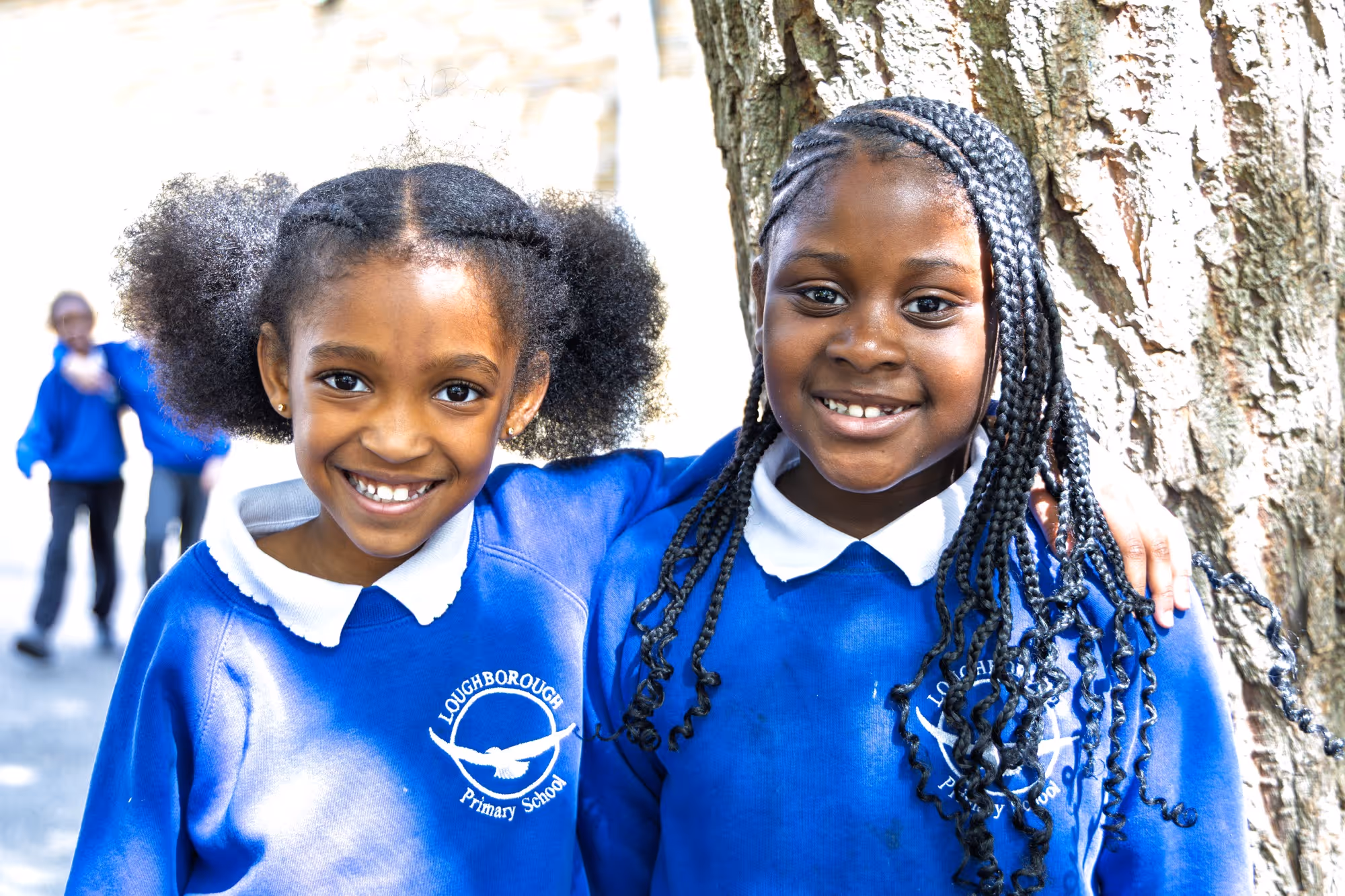 Two smiling girls in blue Loughborough Primary School uniforms standing outdoors by a tree.