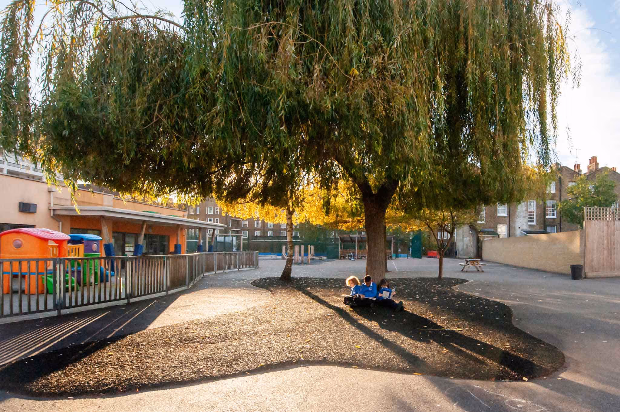 Three children in blue uniforms sitting under a large tree reading books in a school playground.