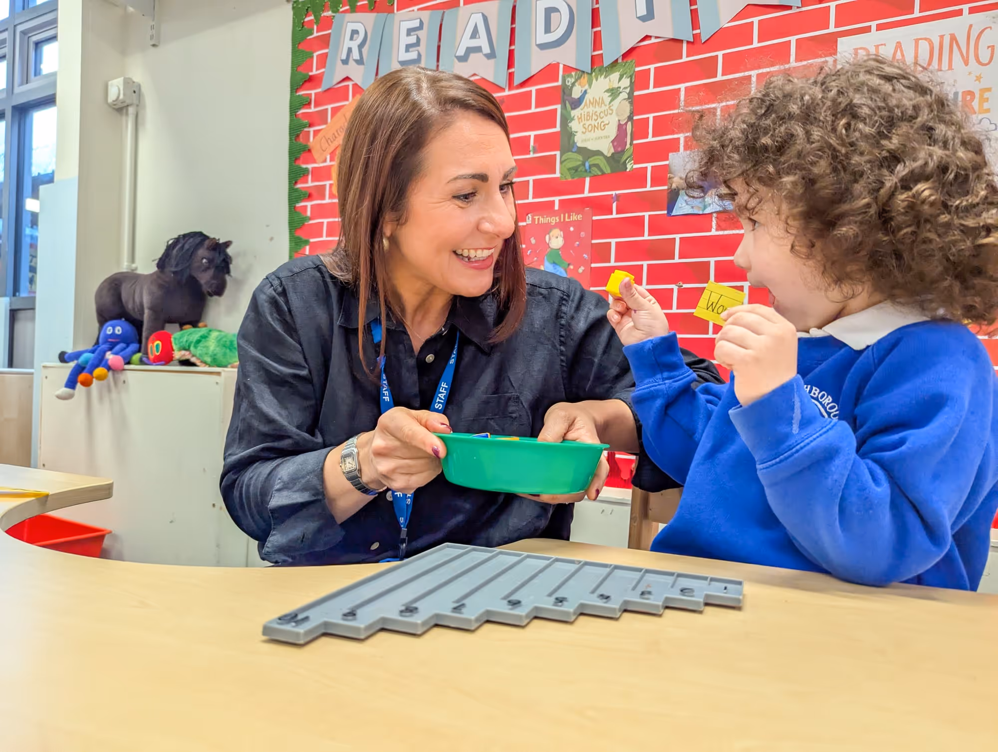 Teacher and child smiling and playing with learning blocks at a classroom table with a reading board behind.