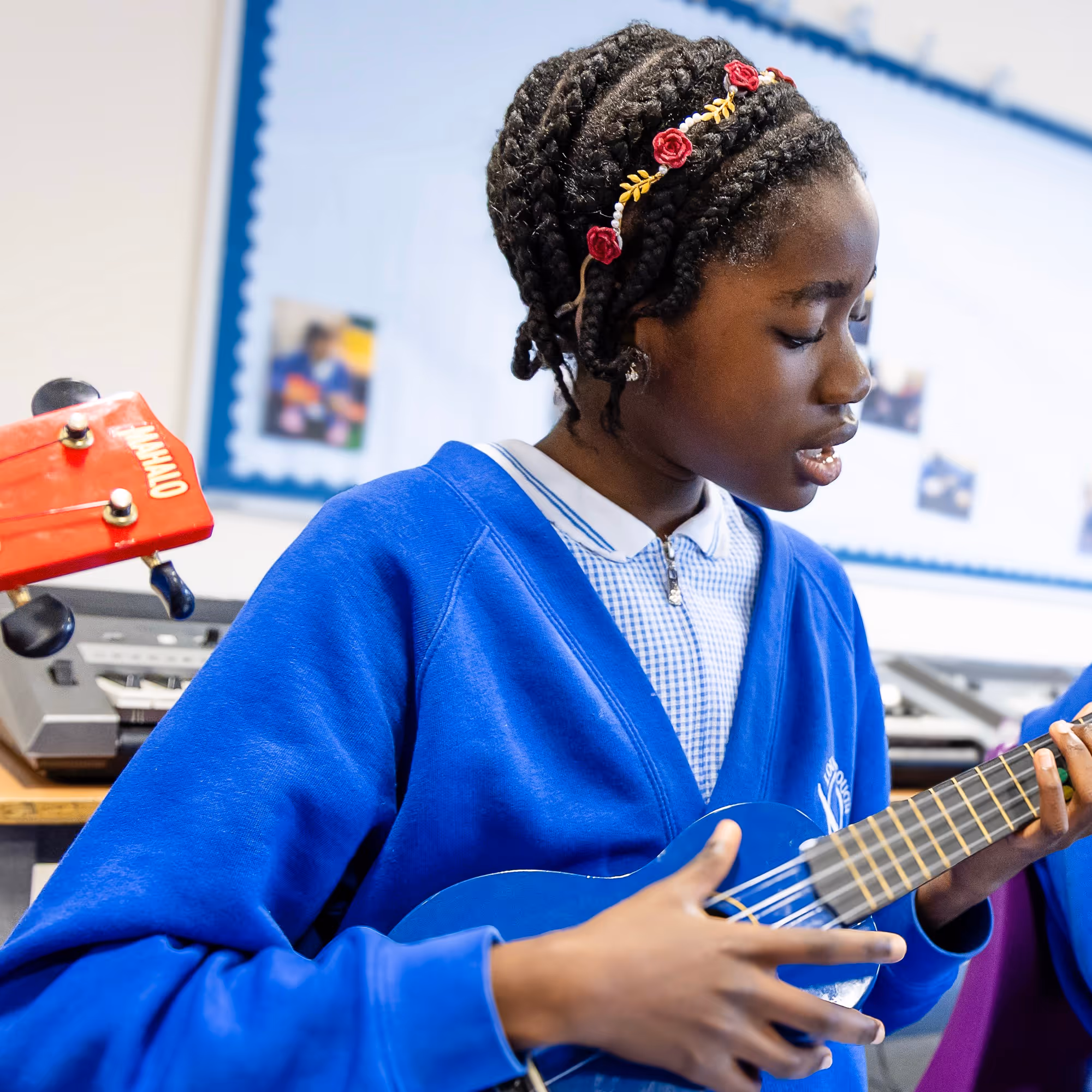 Girl in blue school uniform playing a blue ukulele in a classroom.