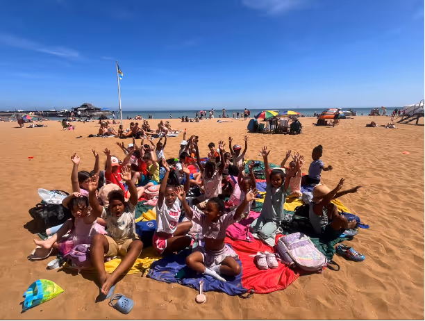 Group of children sitting on colorful blankets on a beach with their hands raised under clear blue sky.