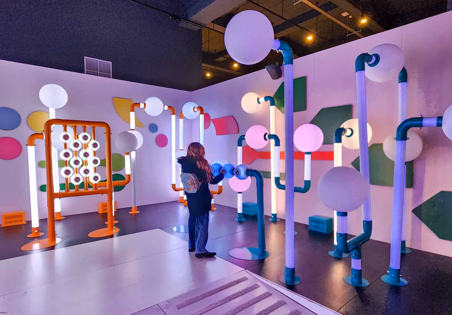 Person interacting with colorful illuminated pipe and sphere art installation in a science museum exhibit.