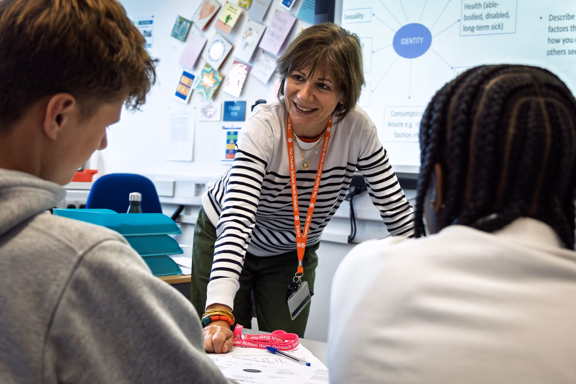 Teacher with striped shirt engaging two students studying a worksheet in a classroom.