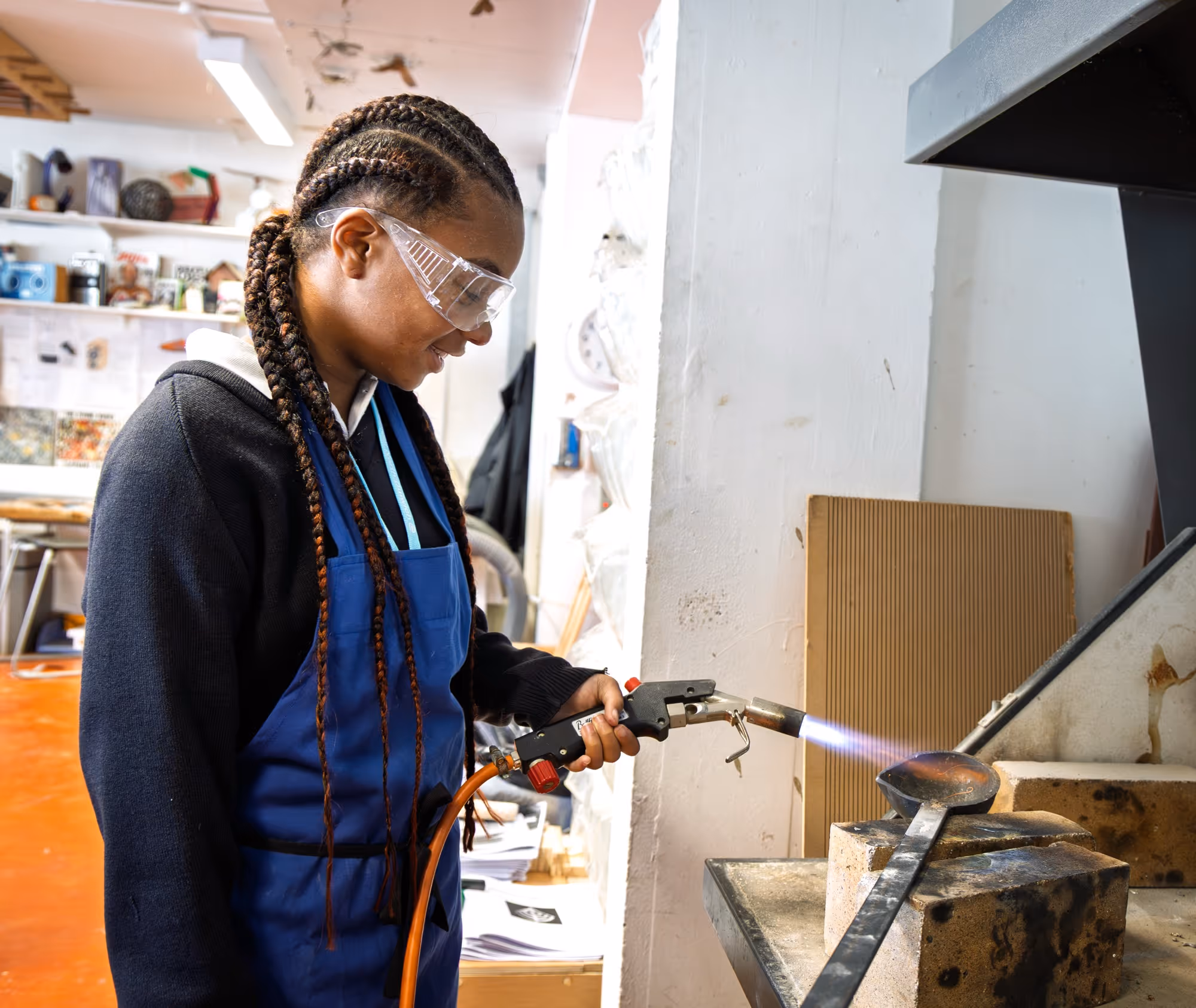 Woman wearing safety glasses and a blue apron using a blowtorch to heat a metal object on a workspace.