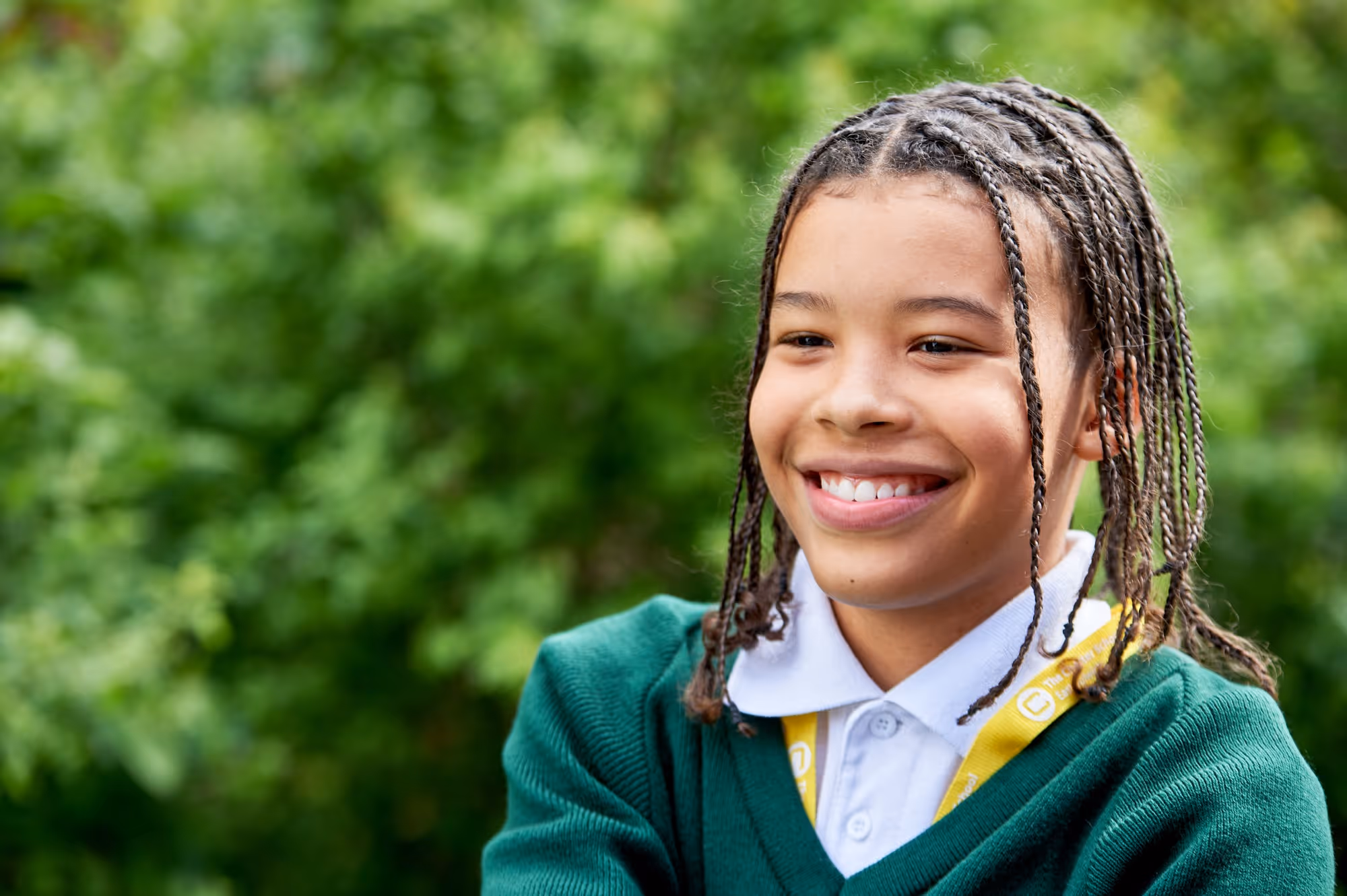 Smiling child with braided hair wearing a green sweater and white shirt outdoors with blurred greenery.