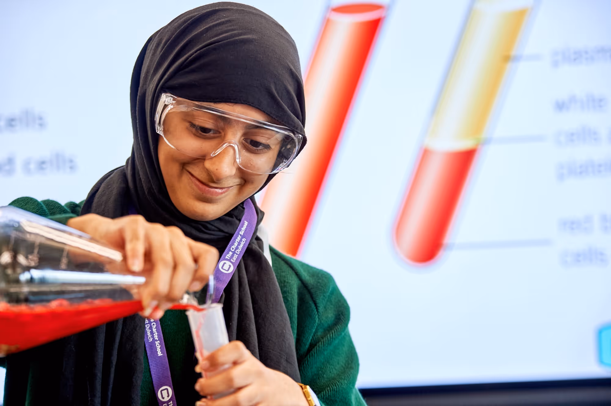 Young woman in safety goggles pours red liquid into a test tube in a science lesson.