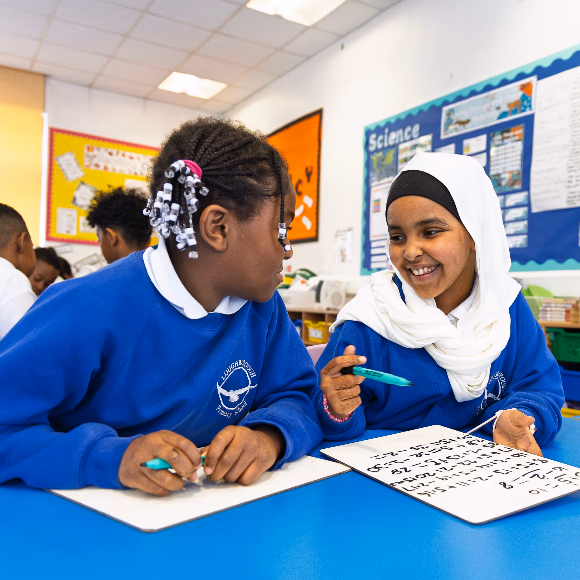 Two schoolgirls in blue uniforms smiling and working on math problems on whiteboards in classroom.