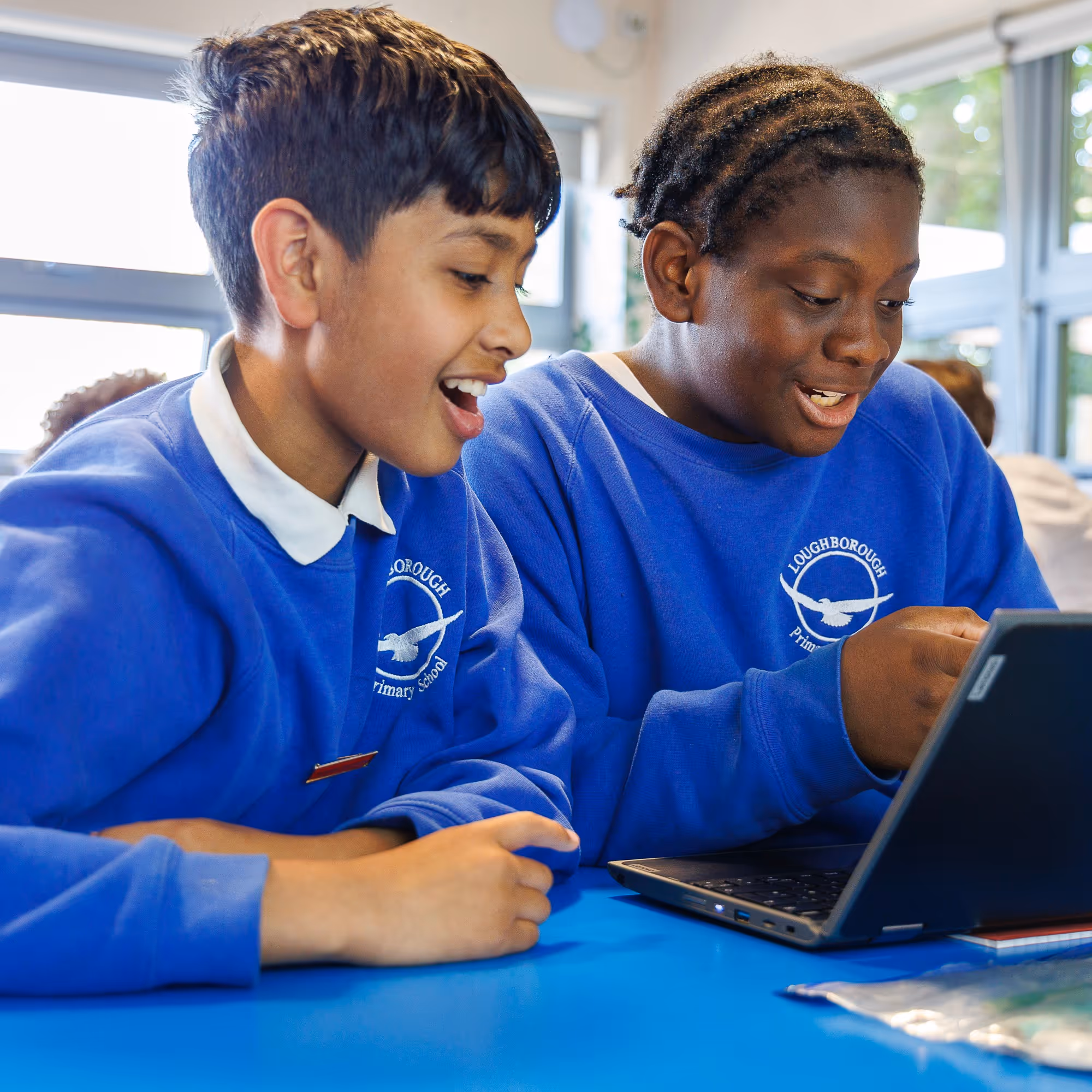 Two boys in blue school sweaters excitedly looking at a laptop screen in a classroom.