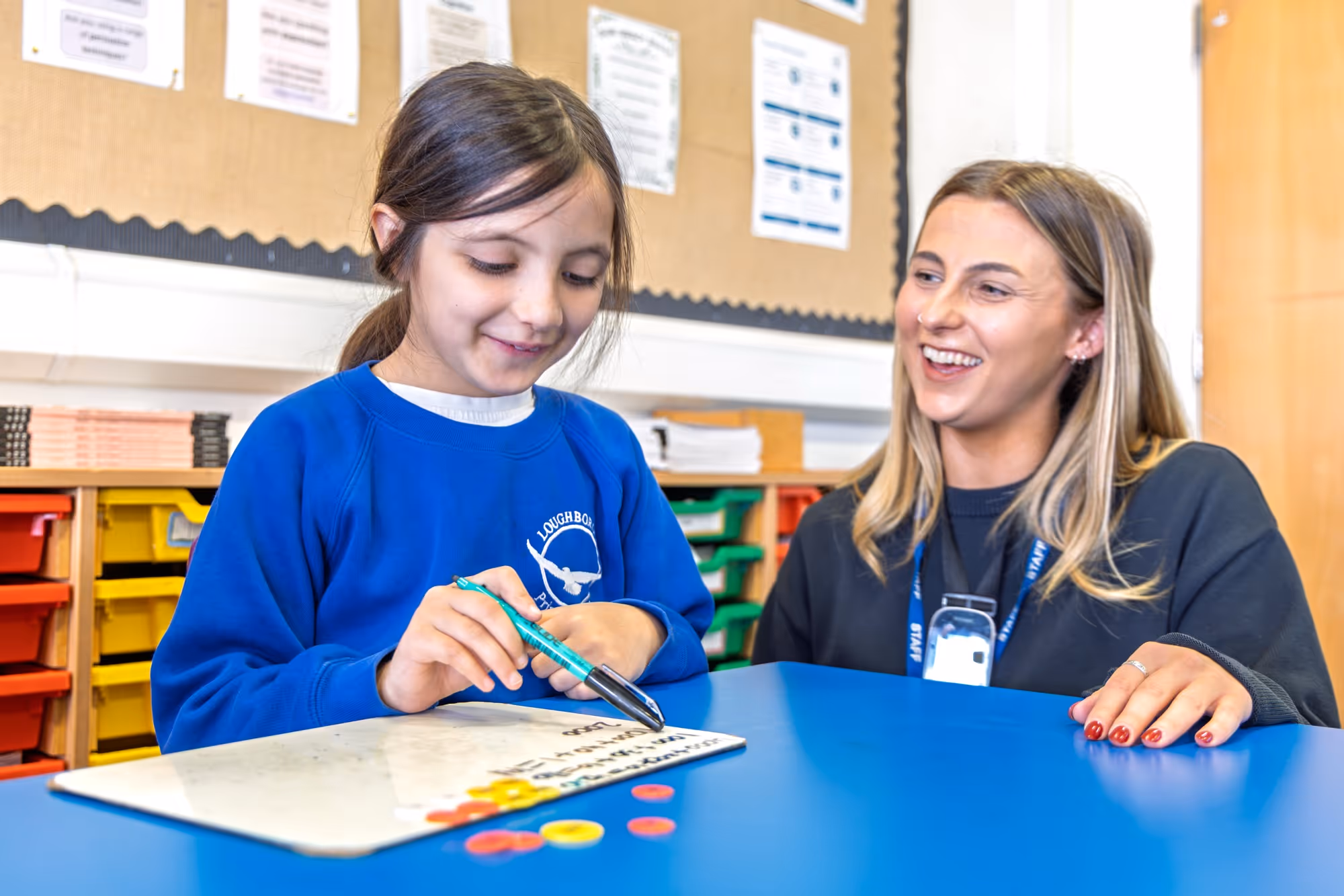 Young girl in blue school uniform writing on whiteboard, smiling woman with lanyard watches her.