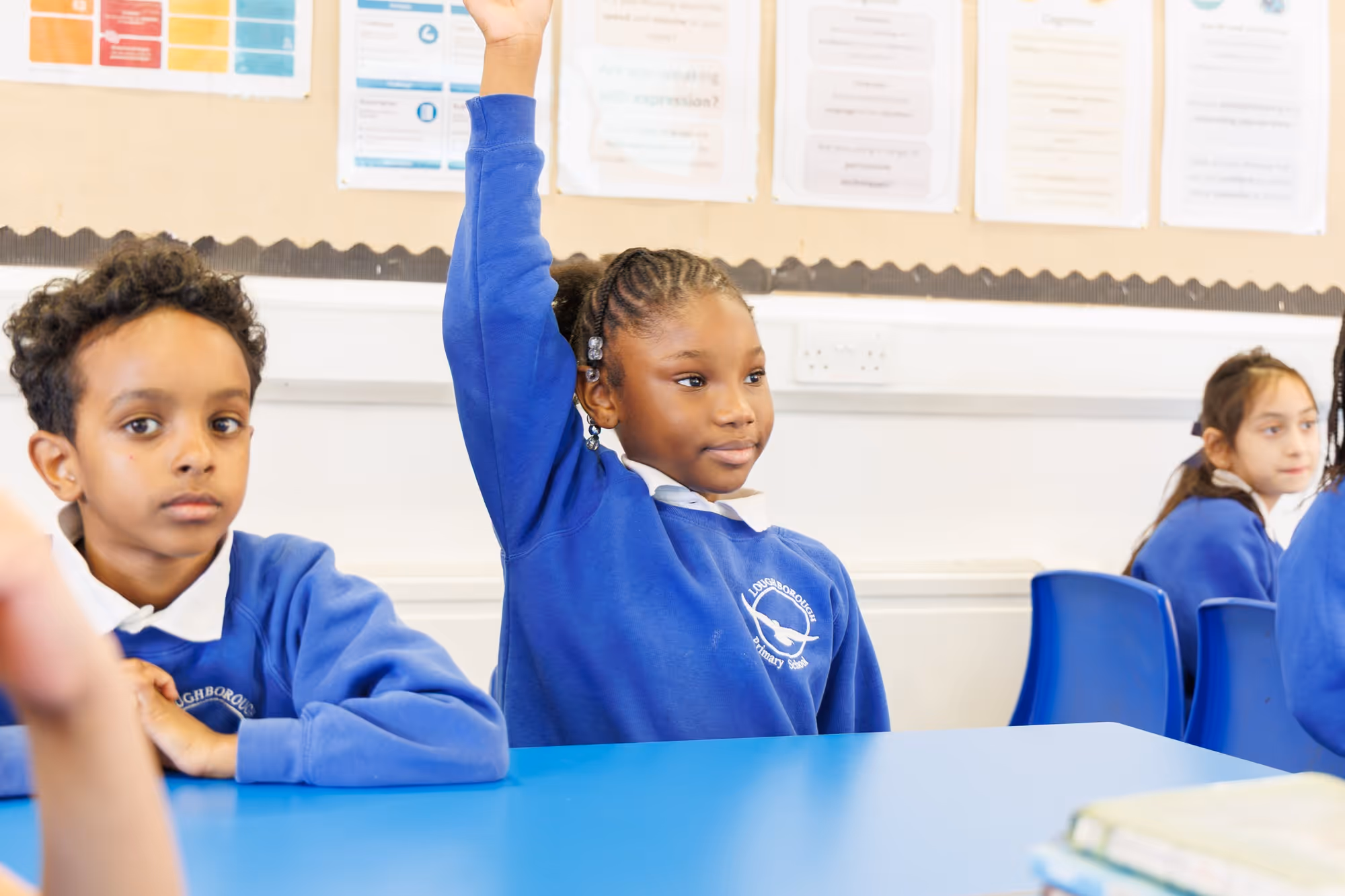 A schoolgirl in a blue uniform raises her hand in class while classmates sit nearby at blue desks.