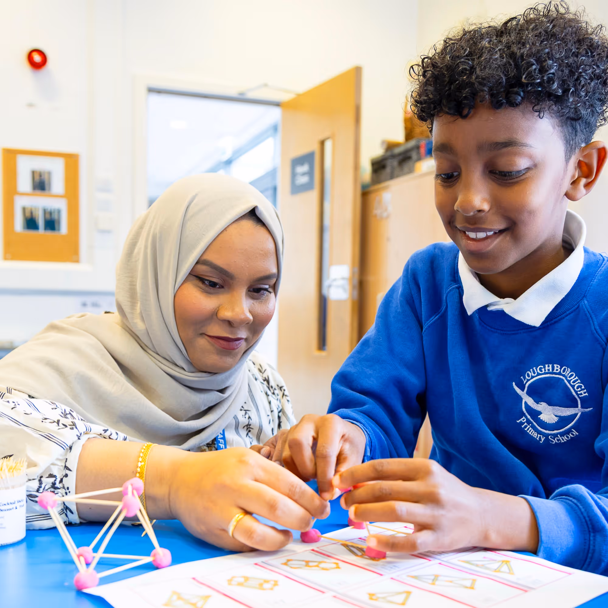 Teacher and student build geometric shapes with sticks and pink connectors at a blue table in a classroom.