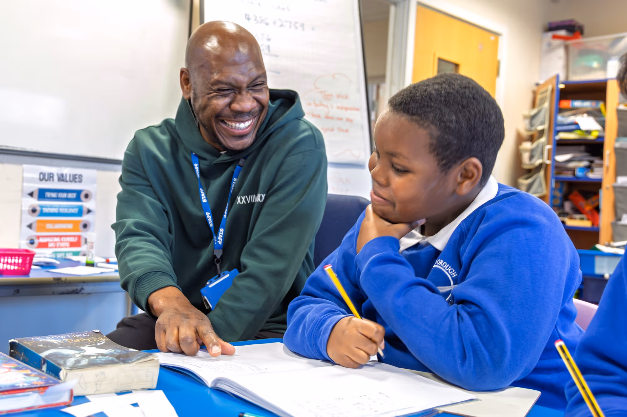 Teacher smiling and pointing at a book while helping a student in a classroom.