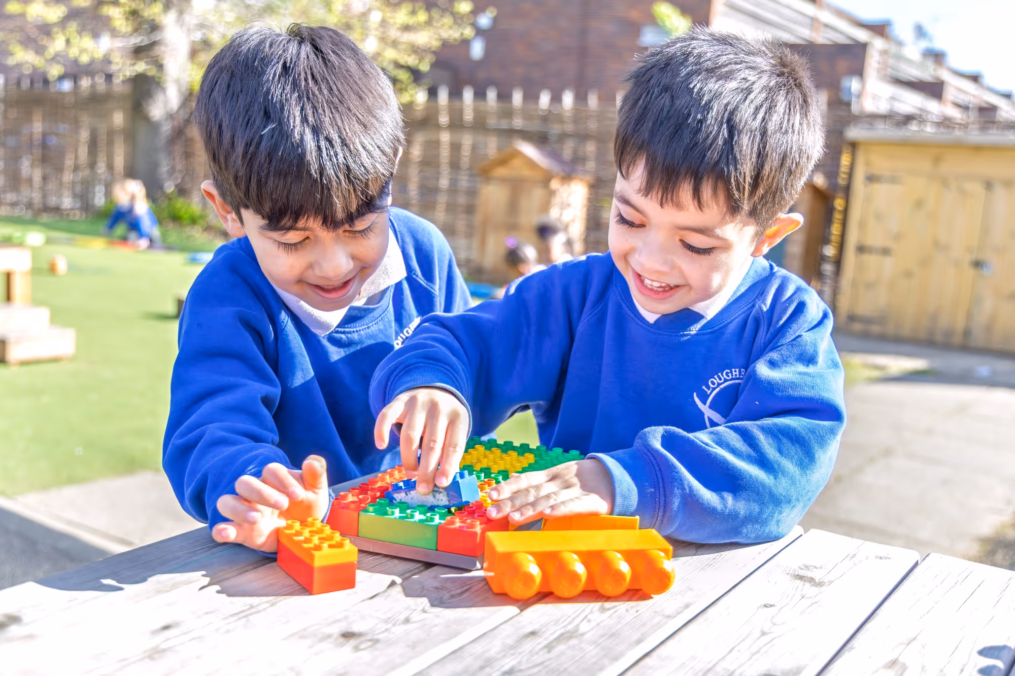 Two young boys in blue sweaters playing with colorful building blocks outdoors on a wooden table.