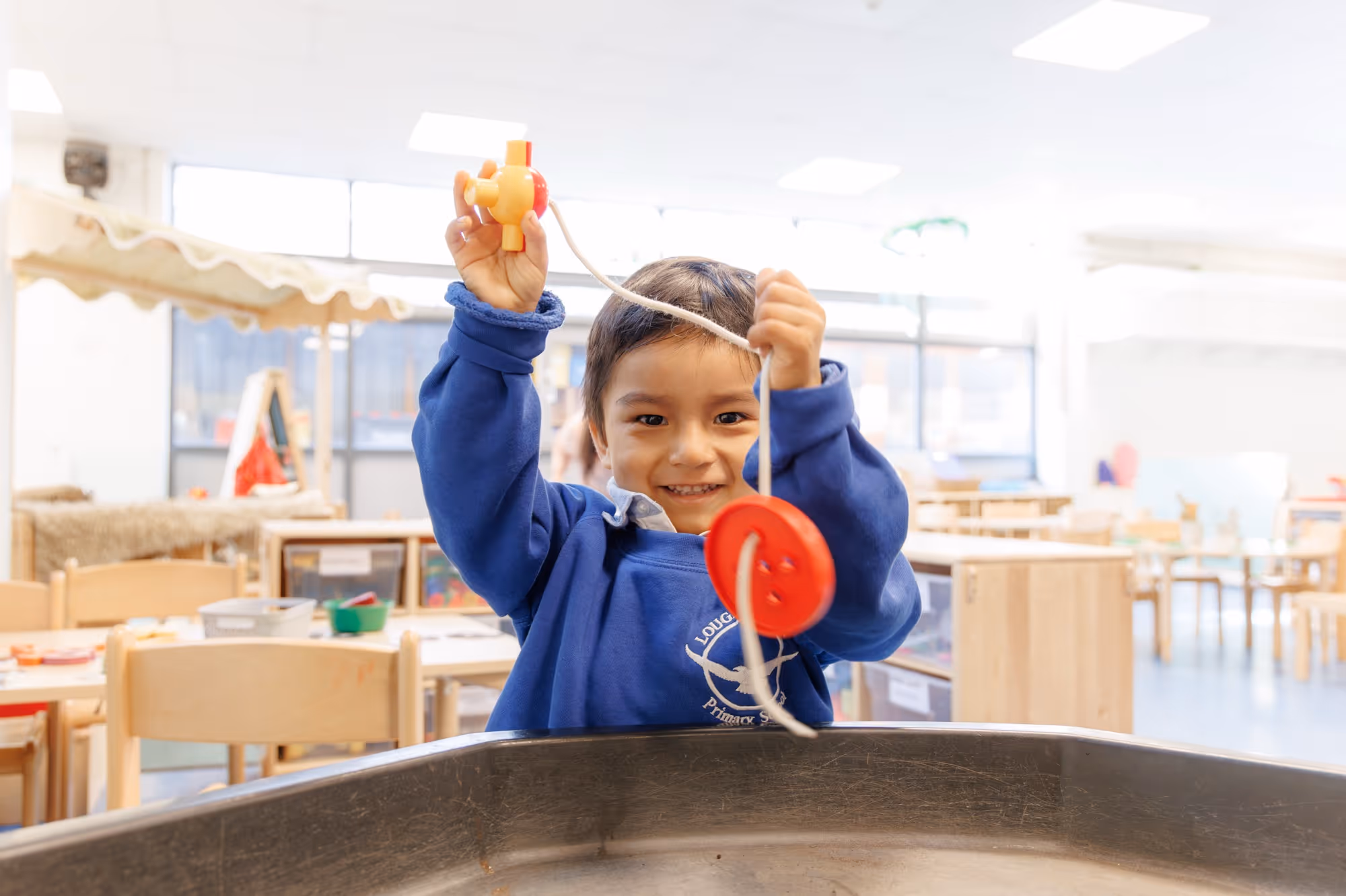 Young child in a blue school sweatshirt holding red and yellow toy connected by a string indoors.