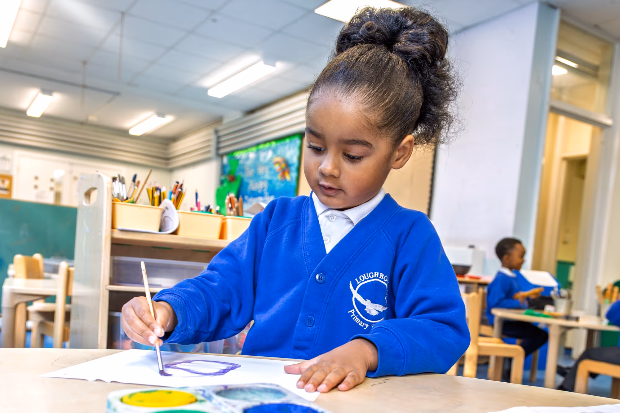 Young girl in blue school uniform painting with brush on paper in a classroom setting.