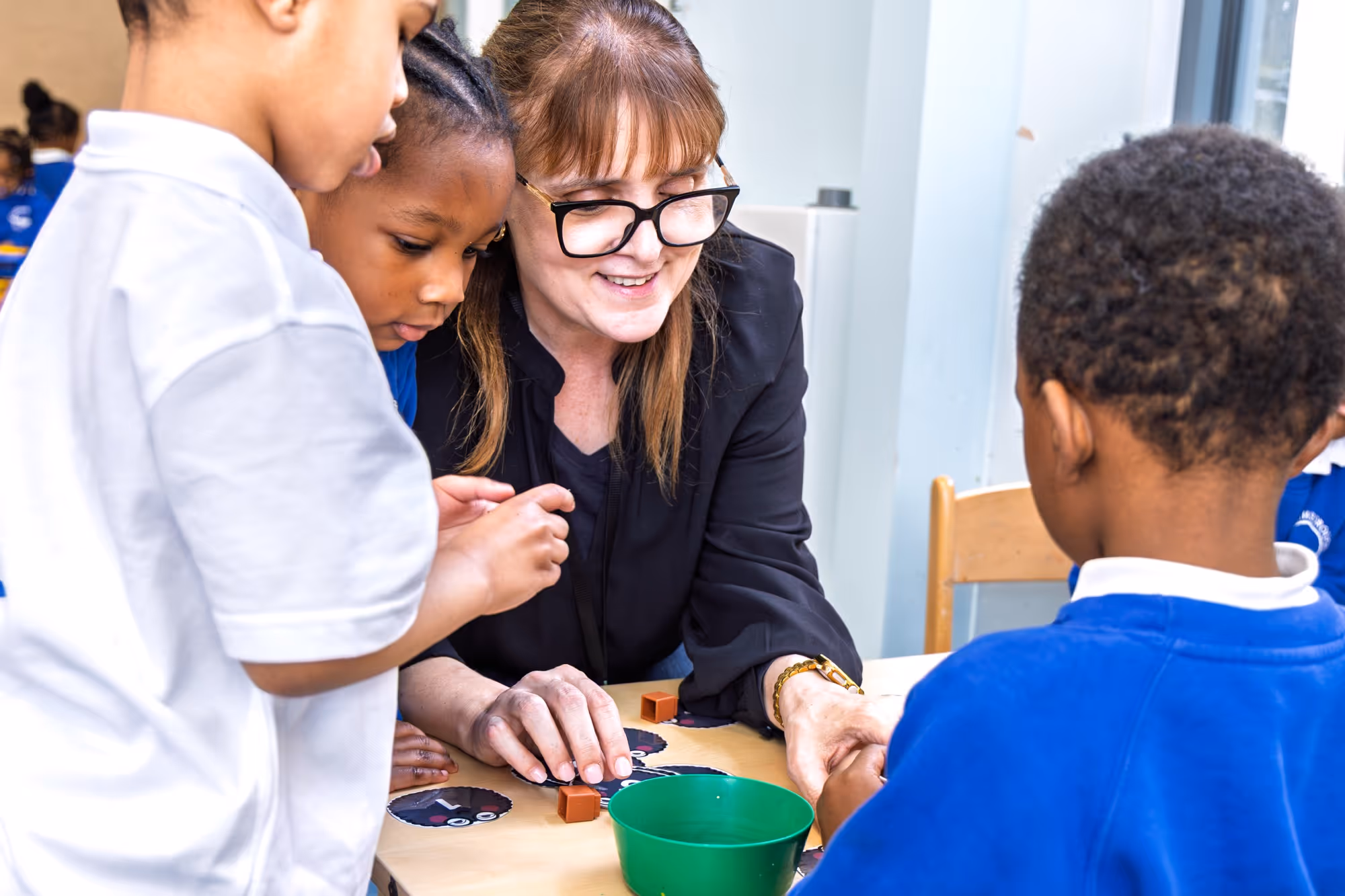Teacher guiding young students at a table with educational cards and blocks in a classroom.