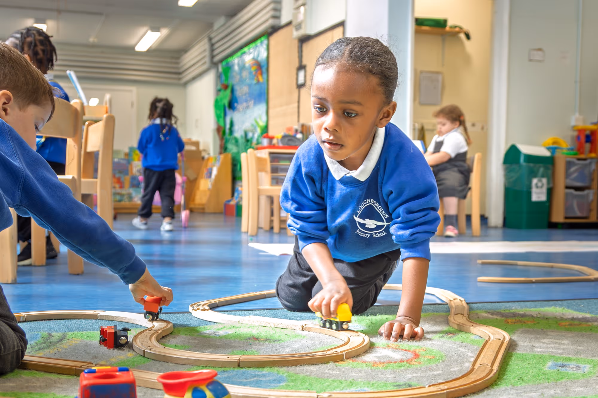 Two children in blue school uniforms playing with a wooden train set on the floor inside a classroom.