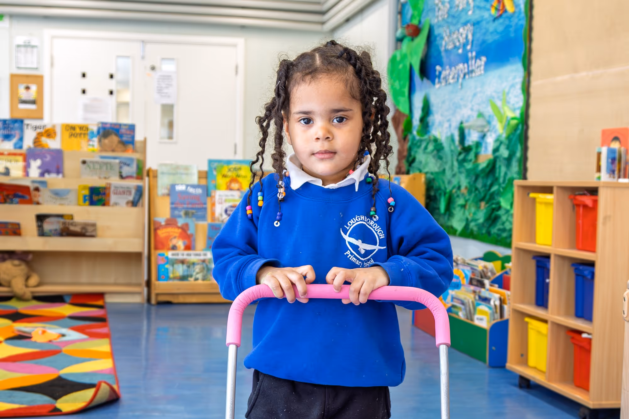 Young girl with braided hair and beads wearing a blue Loughborough Primary School uniform in a colorful classroom.