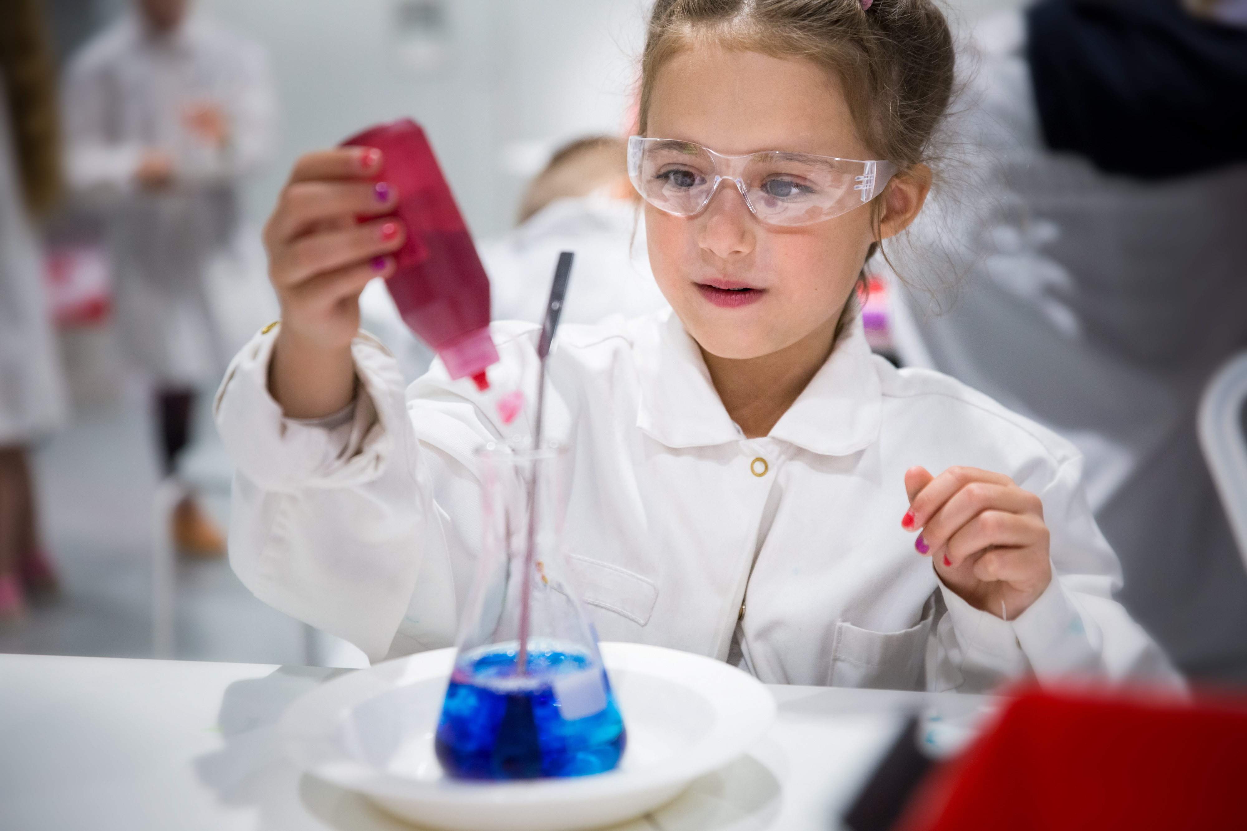 Girl in lab coat and goggles pouring red liquid into a flask with blue liquid during science experiment.