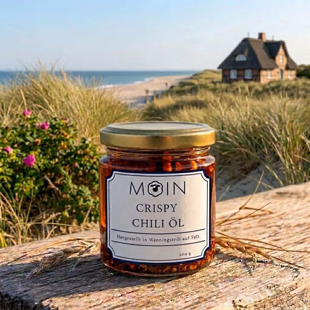 Ein Glas mit knusprigem Chiliöl und Gewürzen auf einem Holzbrett an einem Sandstrand bei Sonnenuntergang.