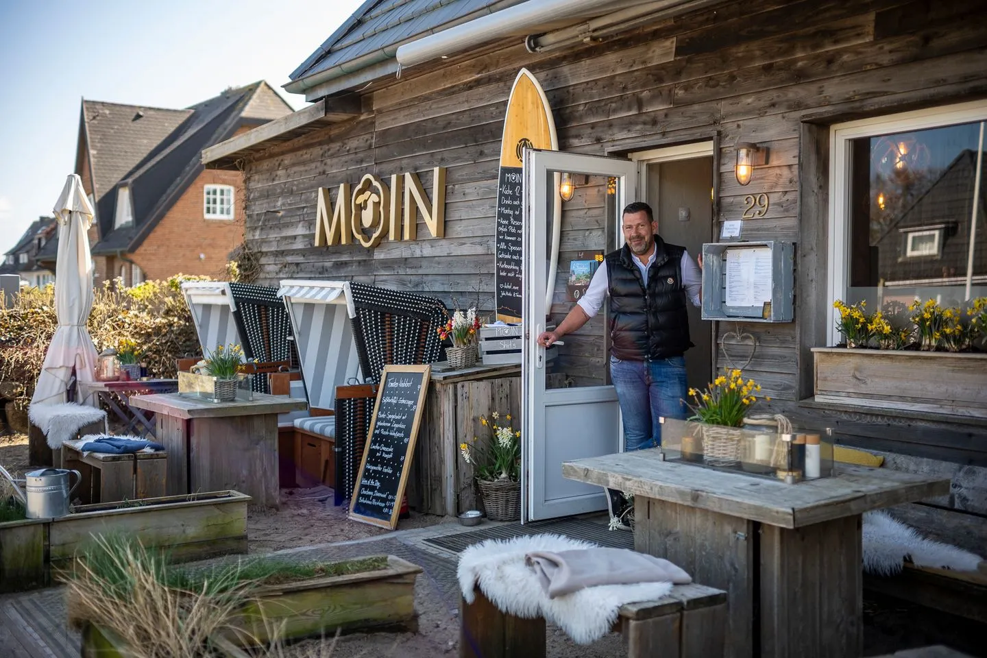 Ein Mann steht im Eingang eines hölzernen Restaurants namens MOIN, das im Freien mit Tischen, Stühlen und Frühlingsblumen bepflanzt ist.