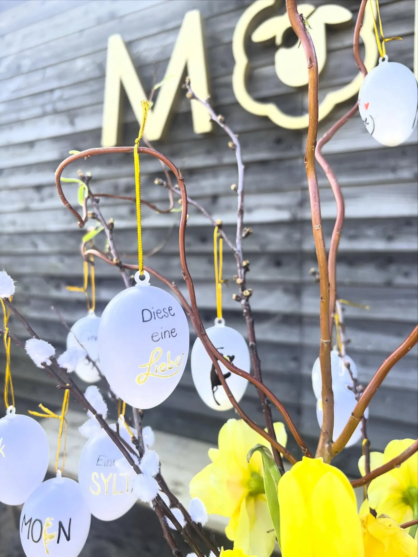 Ein dekorativer Osterbaum mit weißen, ovalen Schmuckanhängern an den Zweigen und leuchtend gelben Blumen am Fuß.