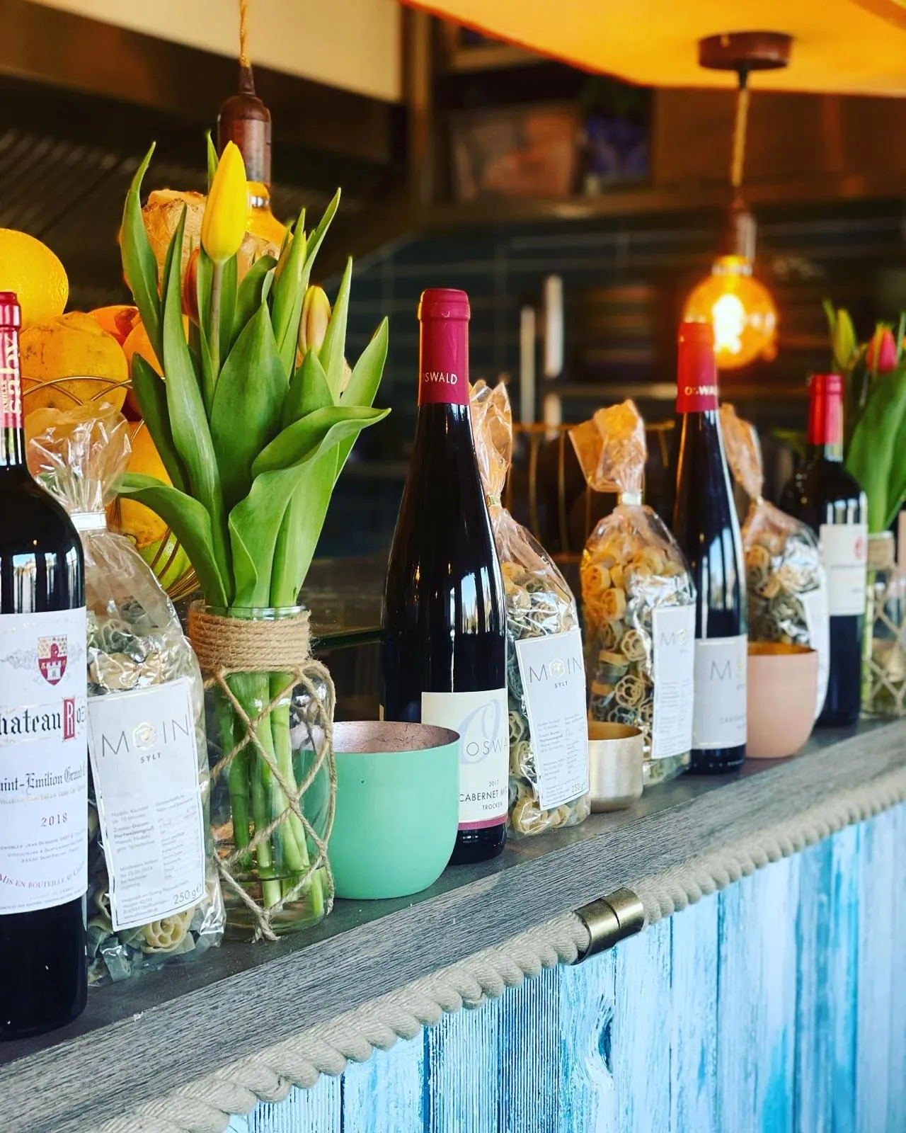 Flaschen Wein, Packungen Pasta und Vasen mit gelben Tulpen auf einer rustikalen Holztheke mit blauer Farbe.