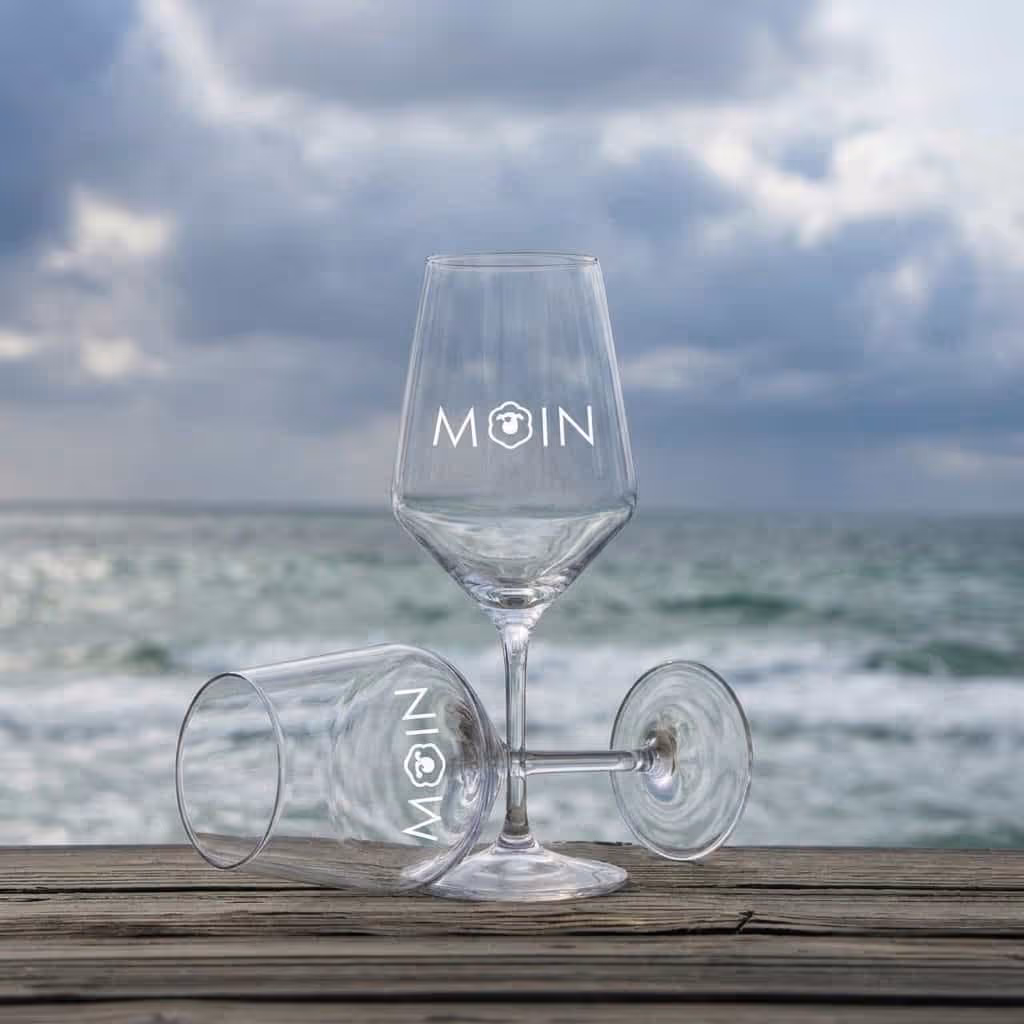 Zwei Weingläser mit Schriftzug MOIN, eins stehend, eins liegend auf Holzplanke vor bewölktem Meer.