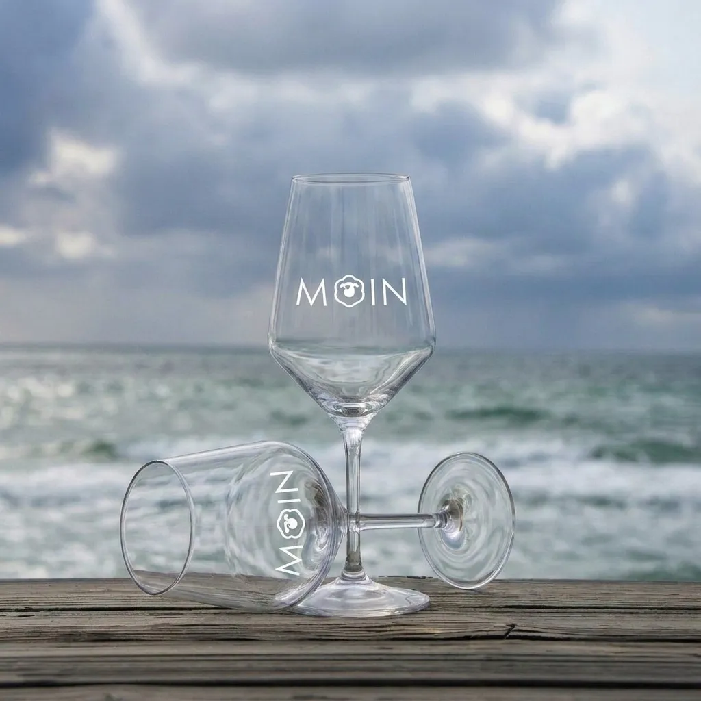 Zwei Weingläser mit Schriftzug MOIN, eins stehend, eins liegend auf Holzplanke vor bewölktem Meer.