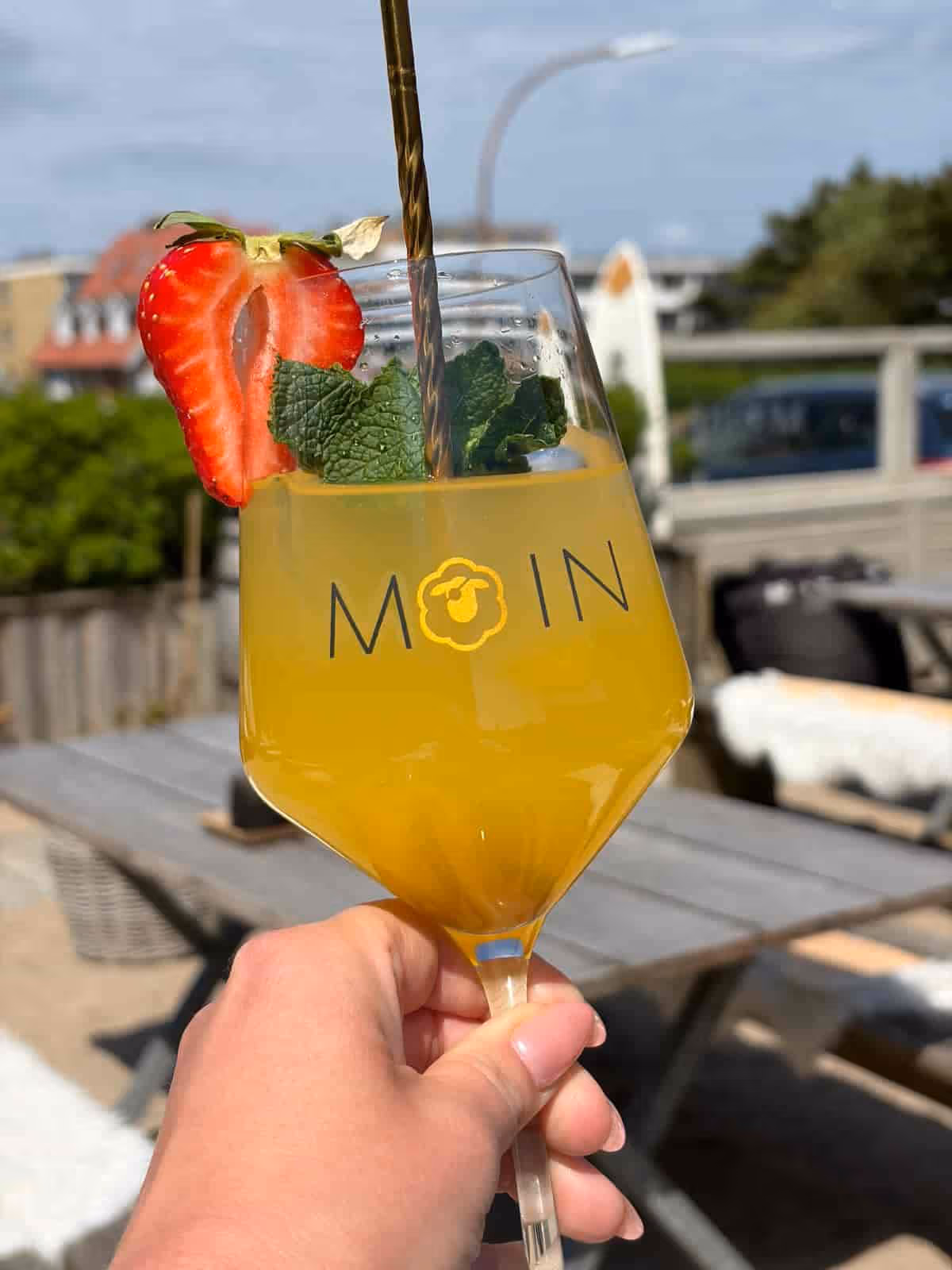 Hand hält ein Glas mit orangem Getränk, Minze und Erdbeere, vor unscharfem Gartenhintergrund.
