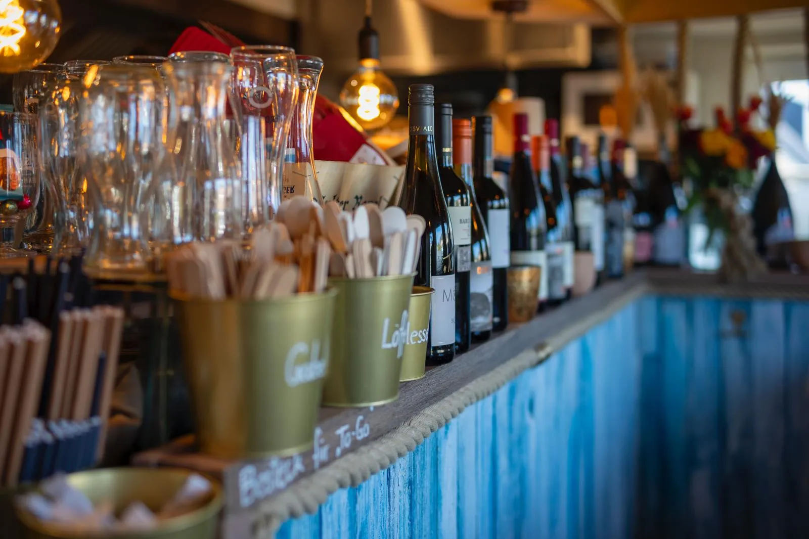 Bar counter with wine bottles, glasses, and containers holding wooden utensils in a cozy setting.